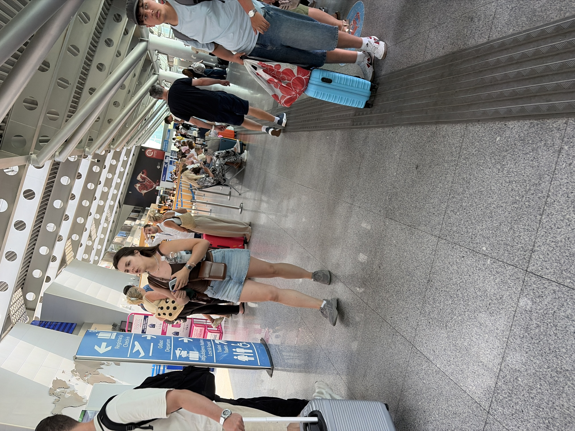 The image depicts an indoor scene, likely an airport or transportation hub, with a modern architectural design featuring a high ceiling with exposed metal beams and circular lights. The floor is a polished gray speckled tile. Several individuals are present, some walking and others seated. In the foreground, a woman in a denim skirt and brown top is holding a smartphone and a water bottle. To her left, a person is walking with a silver suitcase. To the right, a person wearing a light blue t-shirt and denim shorts is holding a red and white floral bag on top of a blue suitcase. A directional signpost in the background indicates "Registration Check-in", "Odlasci Departures", and "Izgubljeno Nađeno Lost & Found". The overall impression is one of transit and travel within a public facility. The location is identified as Golubovci, Montenegro.