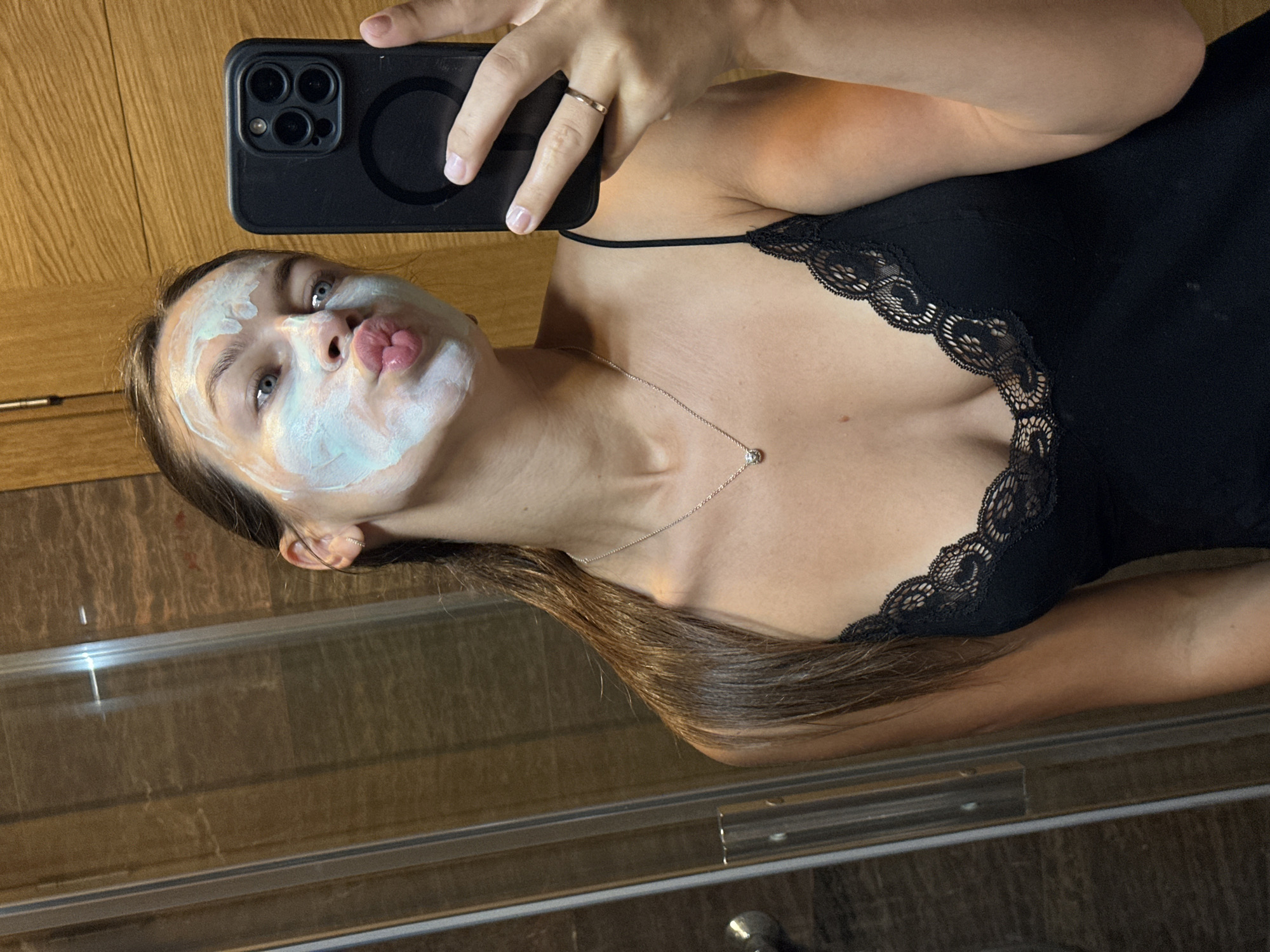 A young woman with brown hair pulled back and a light blue face mask applied to her face is taking a selfie in a bathroom. She is wearing a black camisole with lace trim and a silver necklace with a small pendant. She is holding a black smartphone in her right hand, and her left hand is resting on the phone. The background shows a shower with glass doors and wooden paneling. There is a reflection of the woman in the shower door. The image appears to have been taken in the city of Donji Pobori, Montenegro.