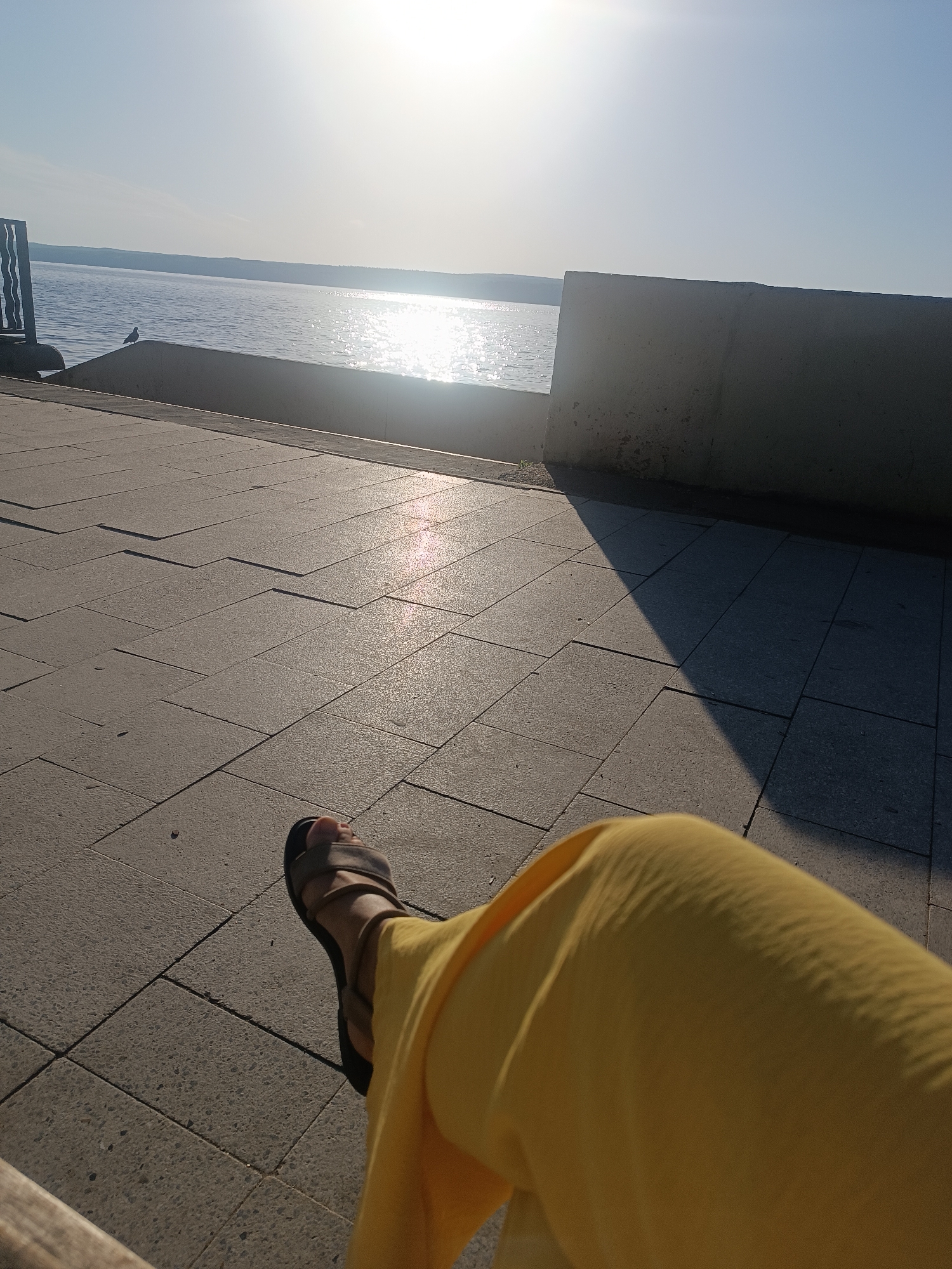 The image shows a view from what appears to be a waterfront promenade in the city of Chaikovsky, Russia. A person is seated, wearing a yellow garment and brown sandals, with their leg extended into the frame. The foreground consists of a tiled walkway, partly in sunlight and partly in shadow cast by a low concrete wall, which runs along the edge of the water. Beyond the wall is a body of water reflecting the sunlight, with a visible horizon. A small bird can be seen perched on the wall in the distance. The sky is bright, suggesting a sunny day. The location seems to be a public space designed for leisure and recreation along the Kama River in Chaikovsky, Russia.