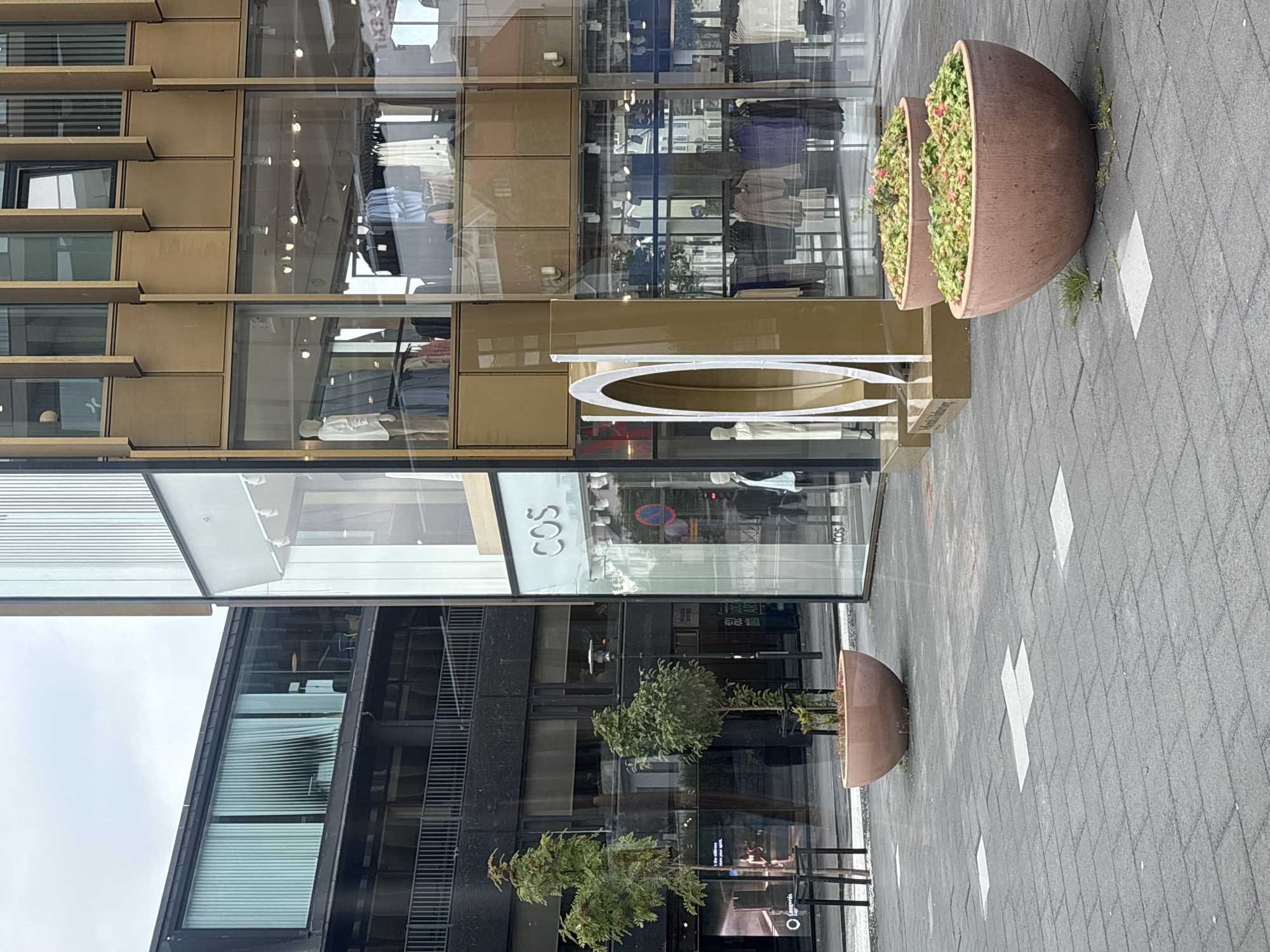 The image shows a street scene in front of a modern building with large glass windows, likely a retail establishment. The building has a gold-colored facade with vertical accents. The storefront displays mannequins and clothing racks filled with apparel. In the foreground, there are two large, round planters filled with green plants and pink flowers. The pavement is made of dark gray tiles. To the left, another building with dark cladding and a metal railing is visible. The sky is overcast. No people are clearly discernible in the scene. This scene is located in Reykjavik, Iceland, on Laugavegur street, a well-known shopping street.