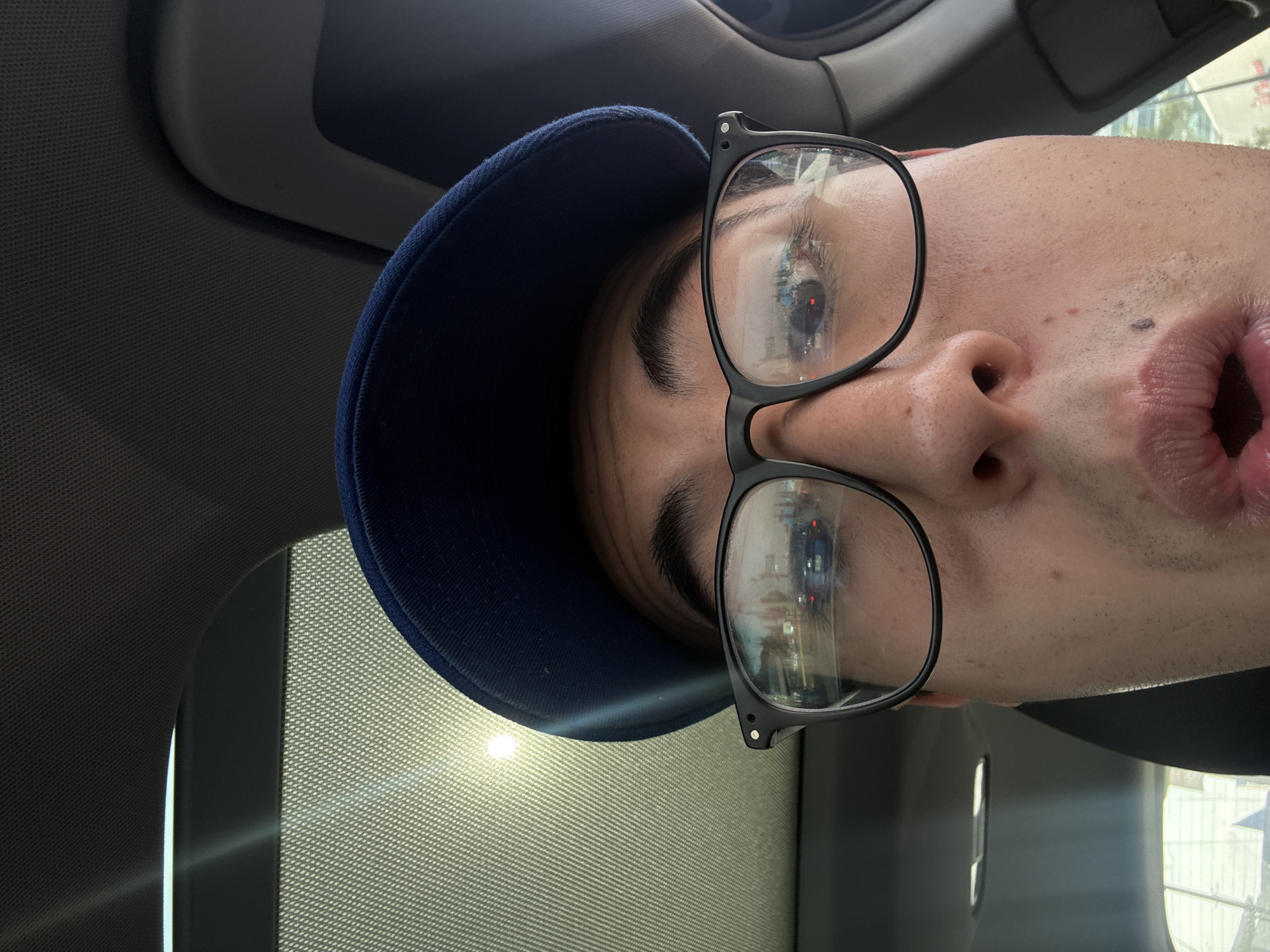 A close-up, vertically oriented selfie captures an individual from the nose upwards, looking directly at the camera. The person's head is tilted to their right, wearing a dark blue baseball cap and black-framed eyeglasses. Their eyes are visible behind the lenses, which reflect an urban scene including buildings, what appears to be a red traffic light, and a bright sky, suggesting the location is a city such as Puebla, Mexico.
The person's mouth is slightly open, perhaps in surprise or about to speak. Faint stubble is visible on their chin and upper lip.
The setting appears to be the interior of a vehicle, possibly a car. The textured grey fabric of the car's headliner is visible in the background, along with a grey grab handle in the upper right corner. A bright source of light, likely sunlight, streams in from the lower left, illuminating the interior and creating a strong reflection. The lighting indicates it is daytime. No discernible text is visible in the image.