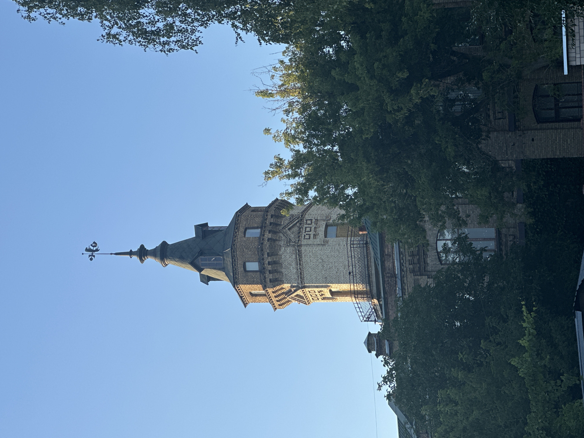 This image captures a detail of the Kyiv Water Tower located in the Podil district of Kyiv, Ukraine, on a clear, sunny day. The tower is constructed from light-colored brick and features a darker, conical roof. At its apex, a weather vane is prominently displayed. The tower is partially obscured by dense green foliage of trees in the foreground and sides. The surrounding buildings, also made of brick, are visible beneath the tower. The sky is a vibrant, cloudless blue, suggesting fair weather.