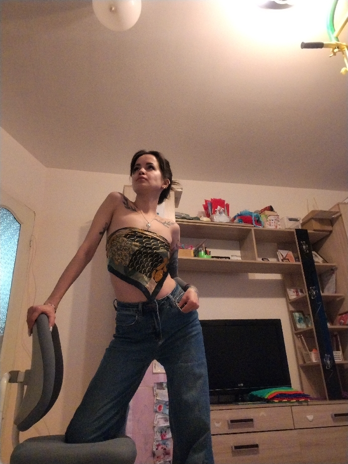 A young woman is captured in an indoor, full-body shot, posing dynamically. She has dark hair styled in a bun or ponytail and fair skin, adorned with numerous tattoos on both arms and shoulders. She is wearing a strapless top fashioned from a patterned silk scarf, featuring dark green, gold, and earthy tones in an intricate, possibly abstract or stylized floral design. Her lower attire consists of high-waisted, wide-leg blue jeans. A delicate necklace with a small pendant is visible around her neck, and she wears earrings.
The woman is positioned with her left knee bent and resting on the grey seat of an office chair, which she leans on with her left hand gripping the top of its backrest. Her right hand is casually placed in her jeans pocket. Her head is tilted slightly upwards and to her left, and her gaze follows this upward direction, giving her a contemplative or confident expression. Her body is angled, showcasing her profile slightly.
The background reveals a lived-in room with light-colored walls and ceiling. A round, white ceiling light fixture is centrally located at the top of the frame. To the right, a multi-tiered wooden shelving unit serves as an entertainment center, featuring a dark-screened television set on a lower shelf. Above and around the TV, the shelves hold various household items, including decorative objects, small boxes, and what appear to be colorful craft supplies or wrapped gifts. To the far right, a tall, narrow, dark panel or decorative piece stands upright, and near the top right corner, a yellow, tubular object hangs from the ceiling, possibly part of exercise equipment or a child's toy. On the left side of the image, part of a door or window with textured or frosted glass is visible. The lighting in the room appears to be artificial, casting an even brightness throughout the scene. No discernible text is visible in the image.