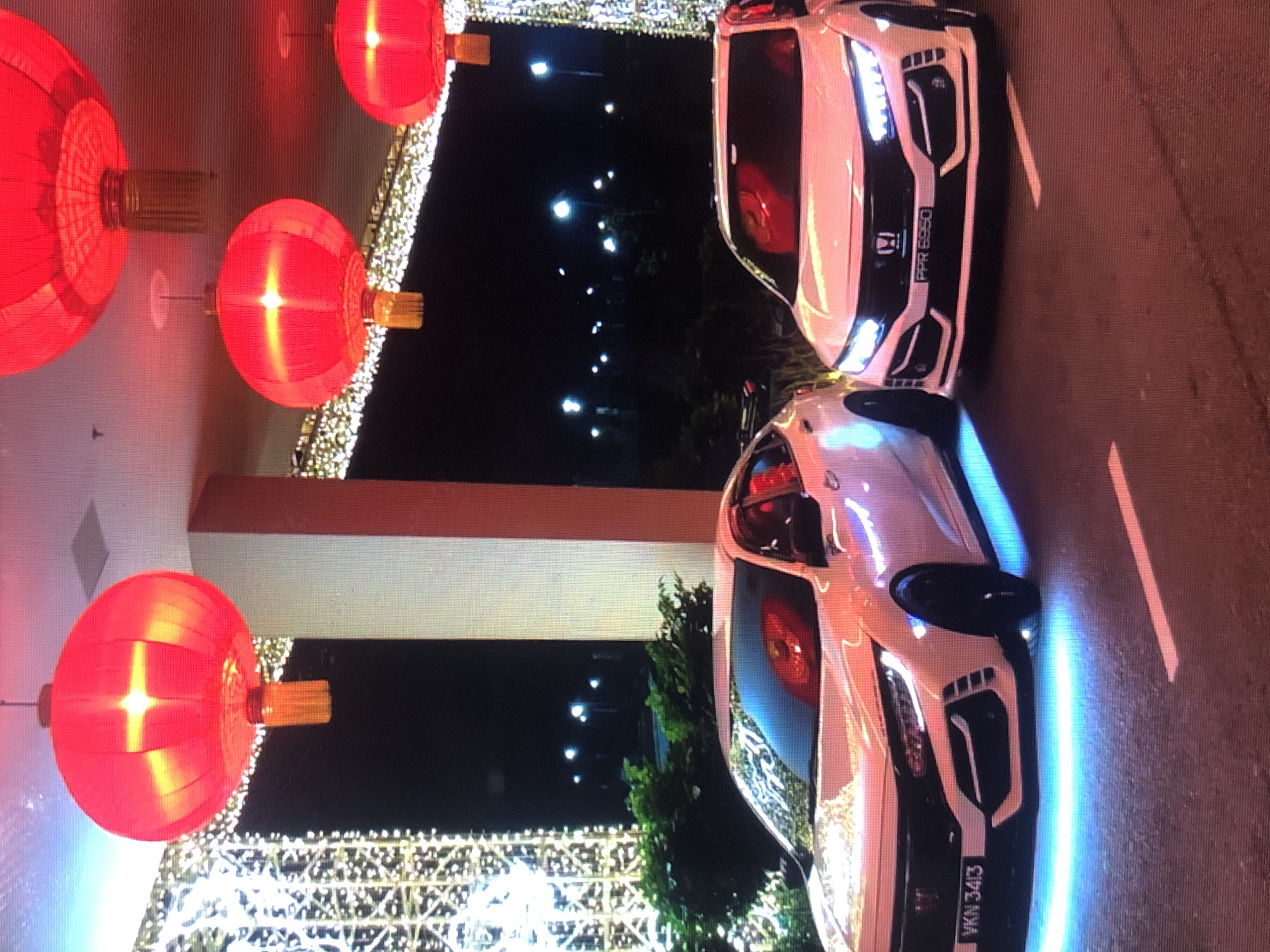 The image shows two white cars parked on a street decorated with strings of lights and red Chinese lanterns. The car on the top has a license plate that reads "PPR 6650". The car on the bottom has a license plate that reads "VKN 3413". The photo was taken at night. There is no evidence to suggest that the image was taken in Lhokseumawe, Indonesia.