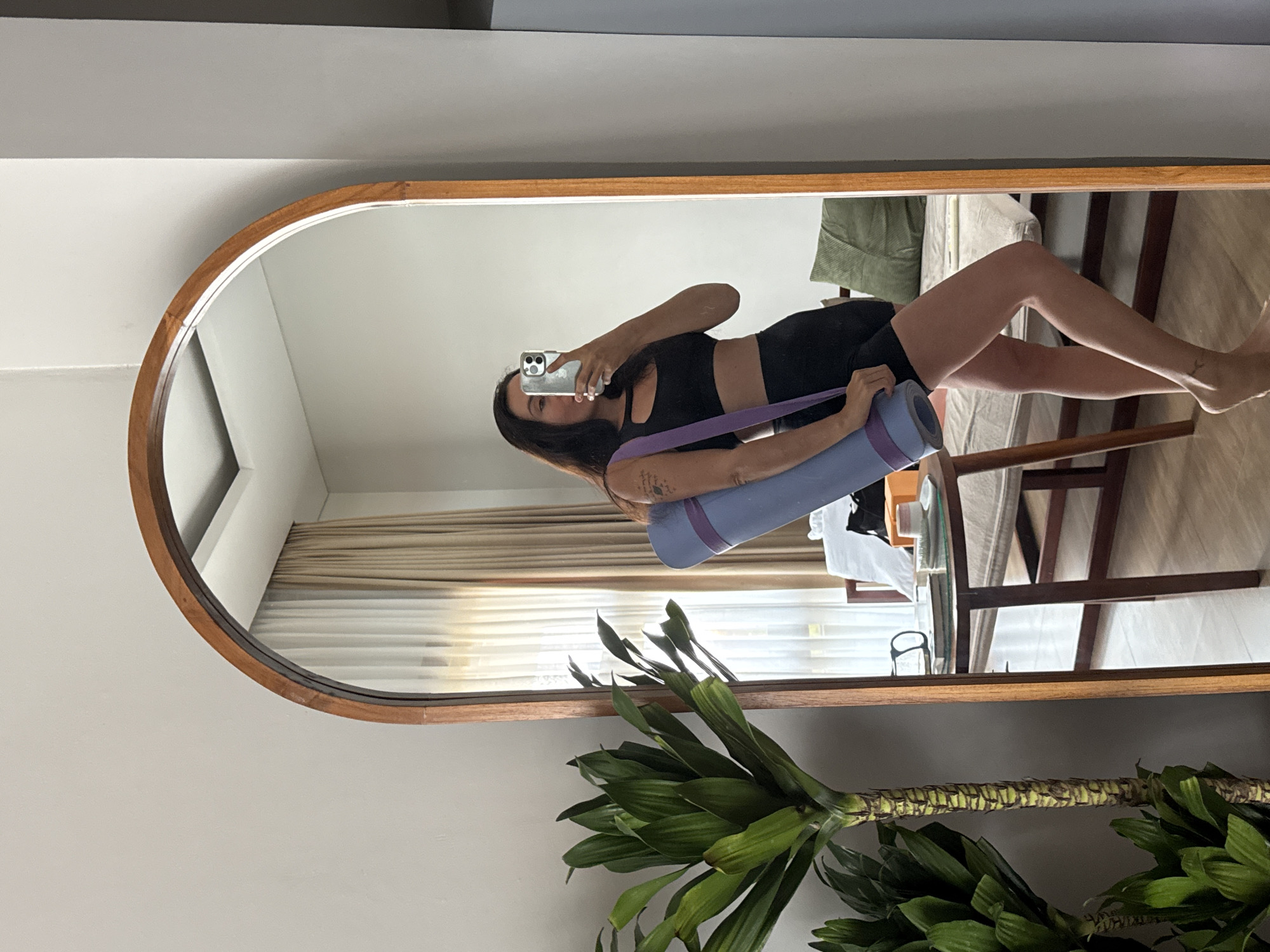 A woman is taking a mirror selfie in what appears to be a bedroom in North Kuta, Indonesia. She is wearing a black sports bra and black shorts and is holding a rolled-up purple yoga mat. The mirror is tall and arched with a wooden frame. In the reflection, a large green plant is visible to the left, and a bed with light-colored linens and a green pillow is in the background. A small wooden table with books and a cup is also reflected in the mirror. The room has beige curtains, suggesting natural light is entering from a window. The flooring is wooden.