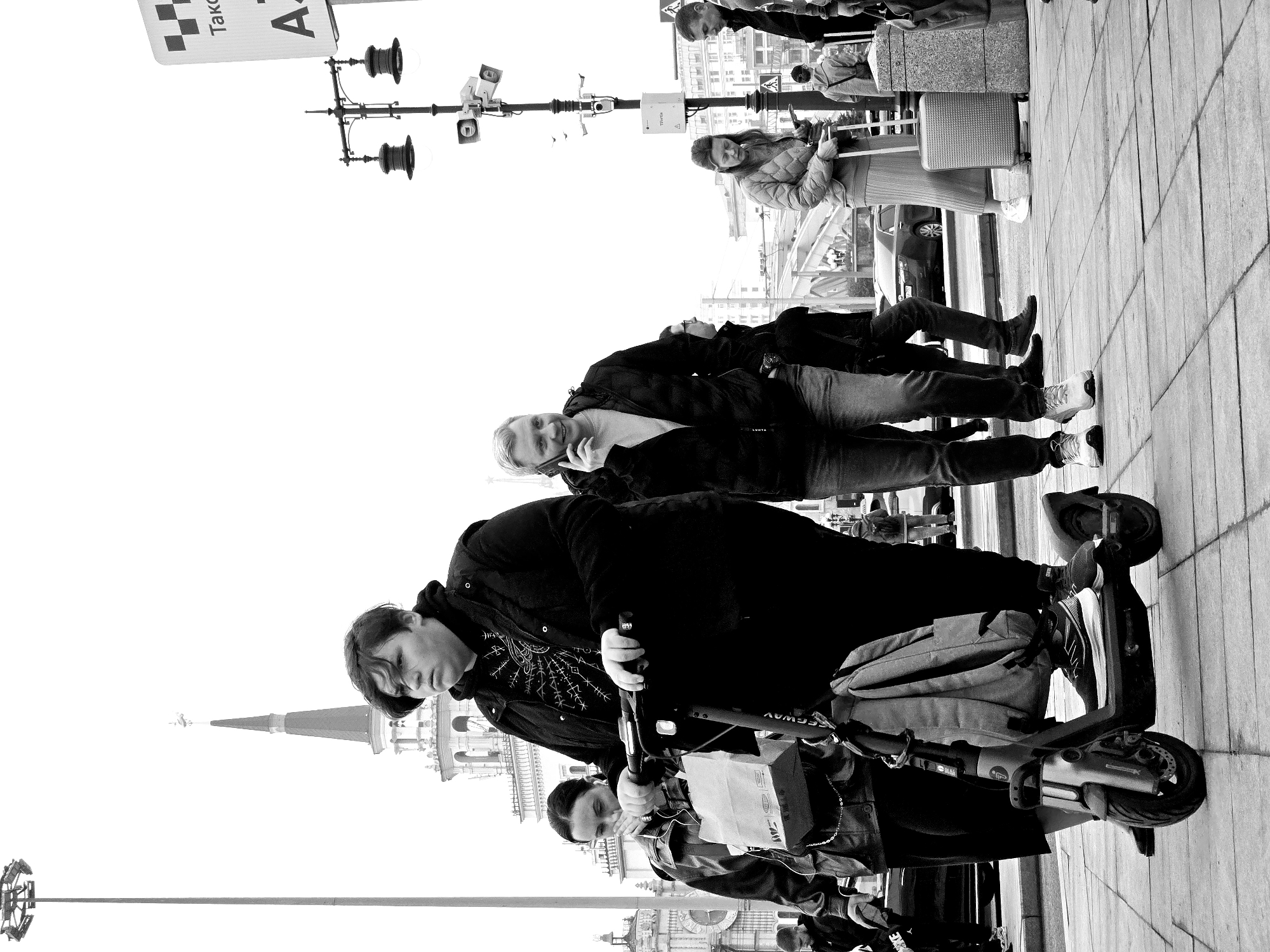 The image, taken in Moscow, Russia, is a black-and-white photograph. In the foreground, a person stands on a Segway scooter with a backpack. Behind them is another person, seemingly also standing on a scooter. In the middle ground, several people are walking and standing. One person is on their phone. A woman is standing by luggage. In the background are buildings, a light pole, and a taxi sign. The environment appears to be an urban street scene.