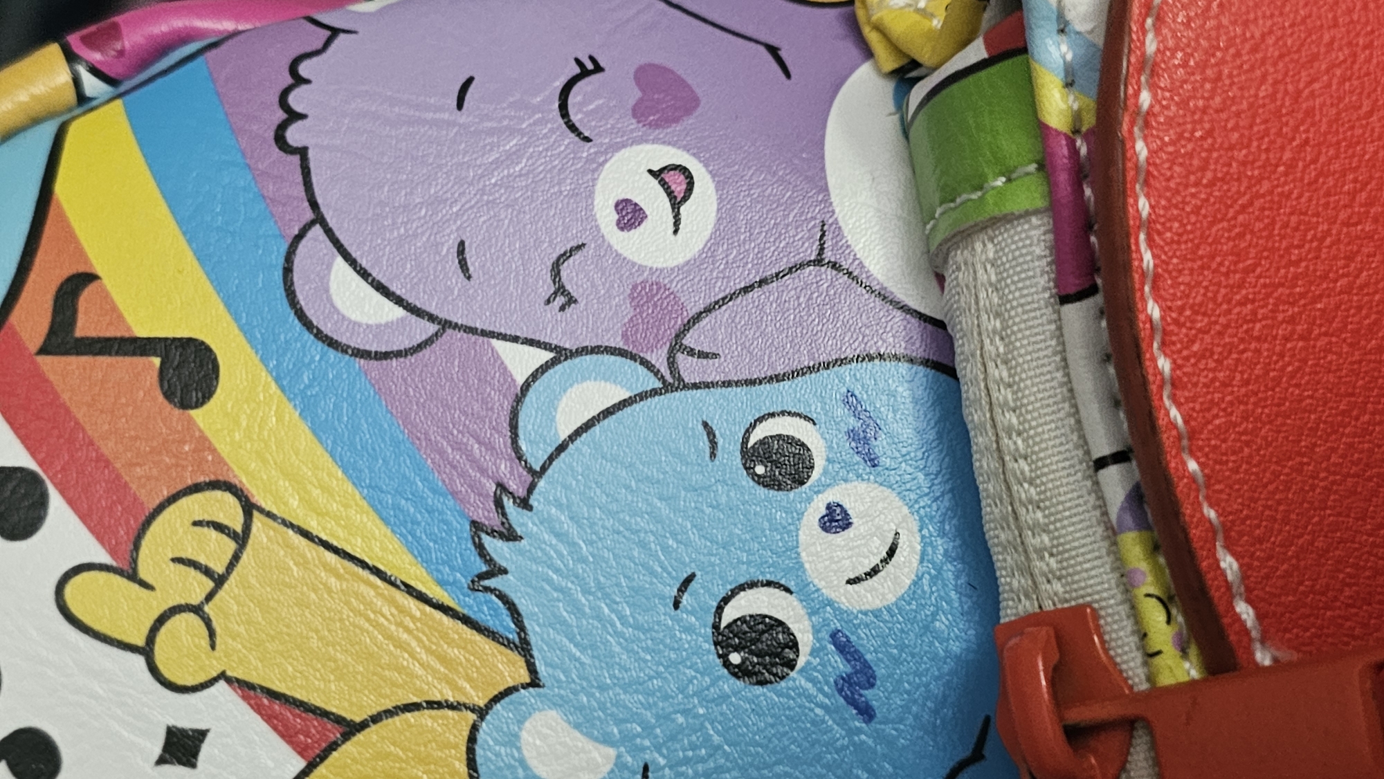 The image shows a close-up of a colorful patterned object, likely a handbag or accessory. The pattern features cartoonish illustrations of Care Bears in shades of blue and purple. A rainbow stripe and musical notes are also part of the design. To the right, there are details of the bag's construction, including a red leather-like edge with white stitching, a gray woven strap, and a red plastic zipper pull.
There is no information in the image or provided to suggest the location is San Bernardino Tlaxcalancingo, Mexico.