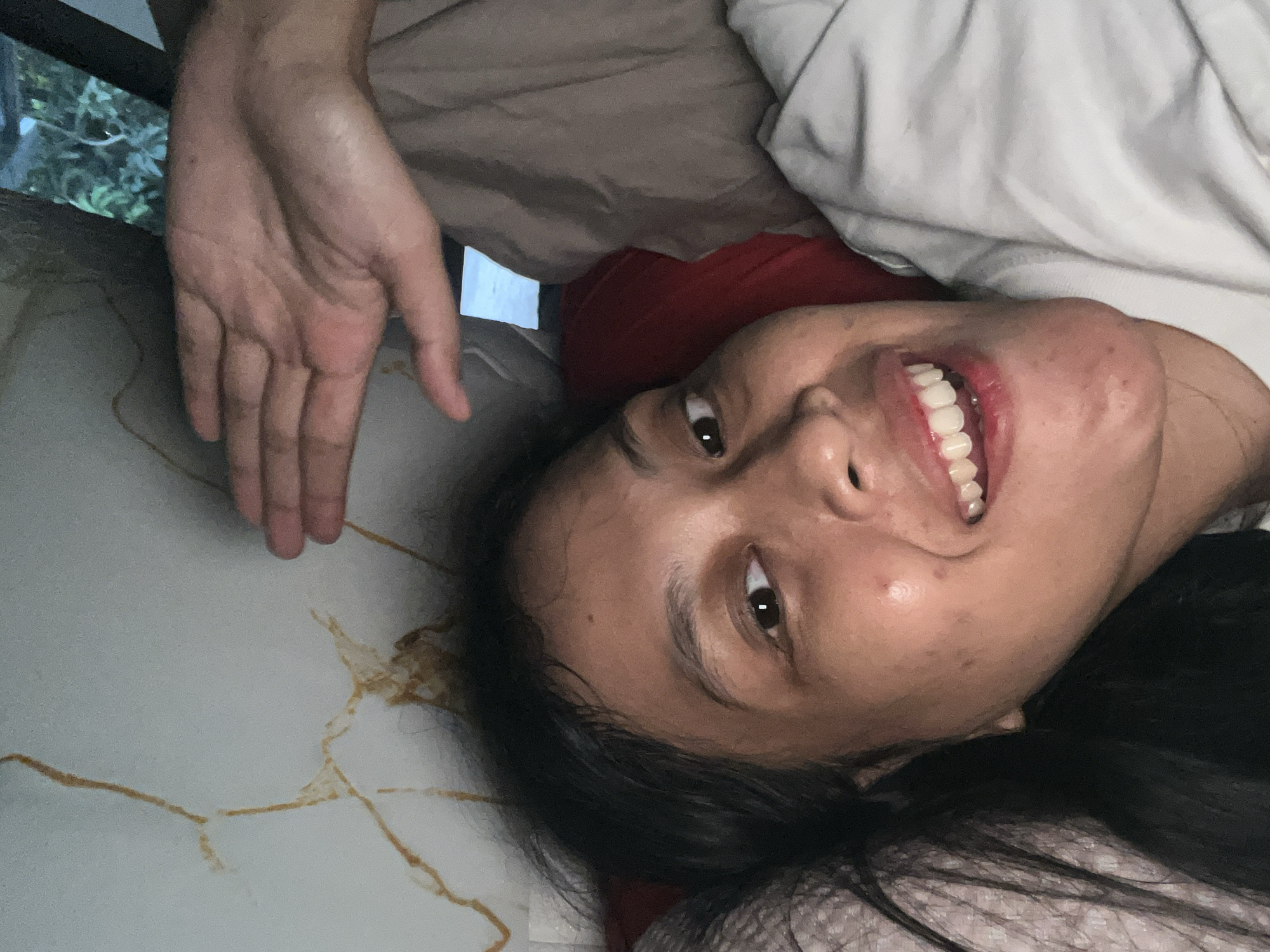 The image shows a person lying down, with their head tilted upwards. They have dark hair and are smiling broadly, showing their teeth. Their eyes are open and looking towards the camera. One of their hands is visible, with fingers spread, positioned above their head. The person is lying on a surface that appears to be a bed, with blankets and pillows around them. The background is out of focus, but suggests an indoor setting with some greenery visible through what could be a window. The lighting suggests it could be daytime. The overall emotion conveyed by the person is cheerful and relaxed. The image is oriented sideways. The location is identified as Kecamatan Pati, Indonesia.