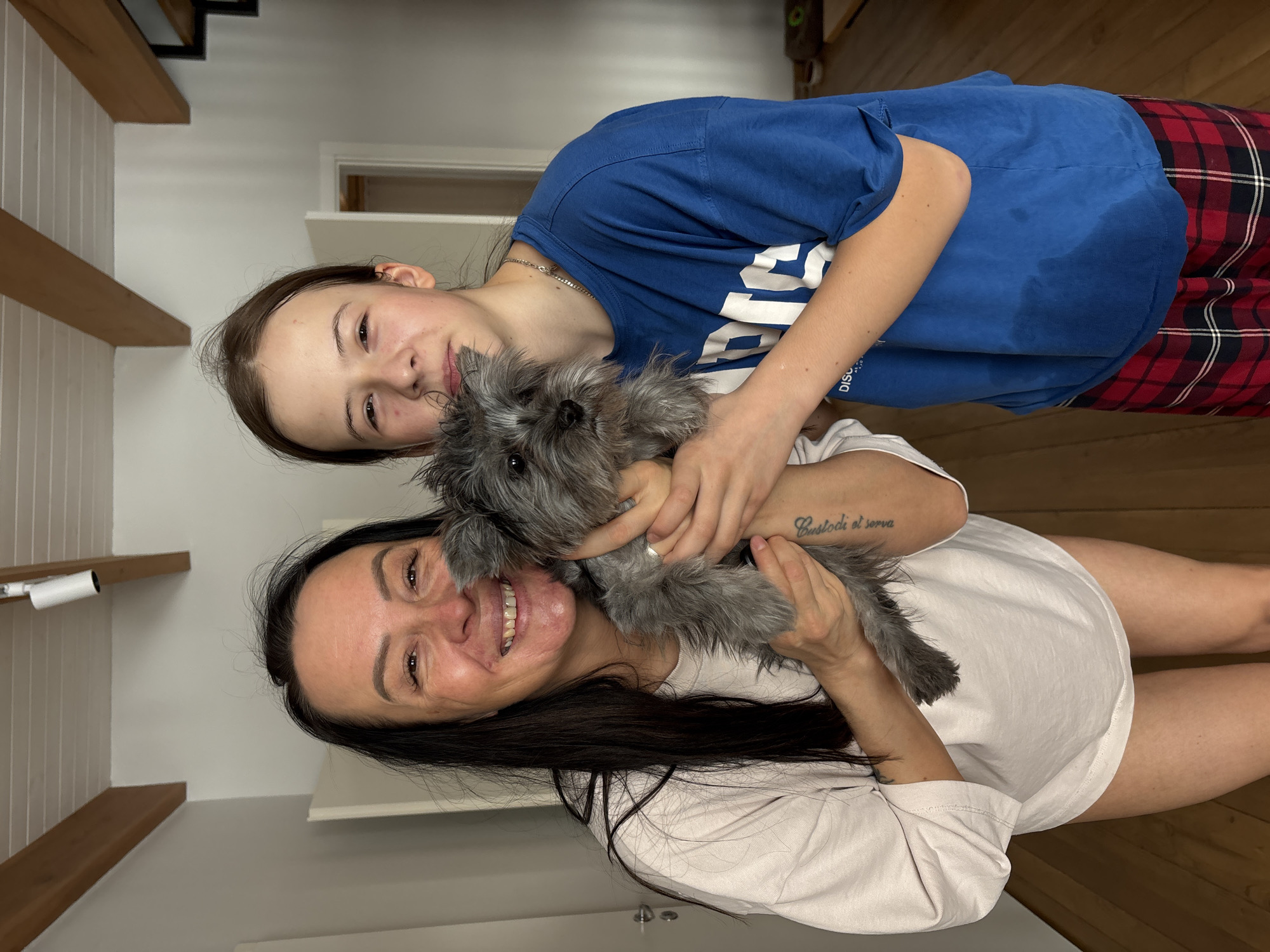 The image shows two females, one older and one younger, holding a gray Schnauzer dog. The older female, with dark hair, is wearing a light-colored T-shirt. She is holding the dog in front of her. The younger female, wearing a blue T-shirt and plaid pants, is standing behind the older female and also has her arms around the dog. They are both smiling.
The background includes a white wall with wooden beams, a doorway, and a wooden floor. The image was likely taken indoors in a home. The location, Chaikovsky, Russia, is not discernible from the image.