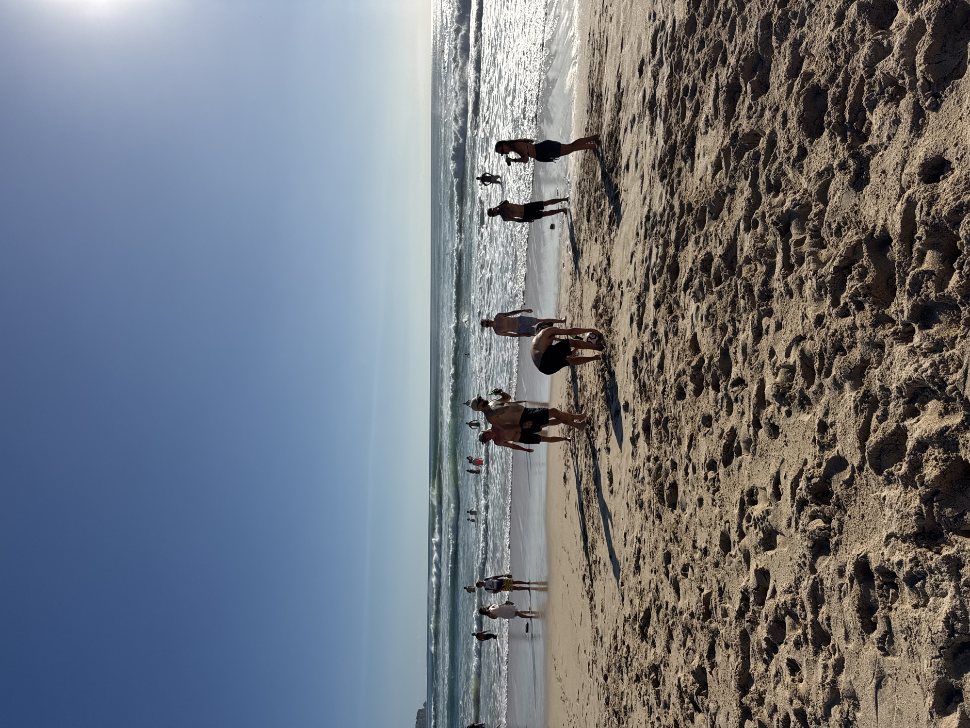 The image shows a sunny day at a beach, likely in Kuta Selatan, Indonesia, with clear blue skies and a bright sun in the upper right corner. Waves are rolling onto the sandy shore where several people are engaged in various activities. In the foreground, a group of individuals are on the sand. One person is bent over, appearing to interact with a soccer ball. Others are standing, some looking out at the water. Further back, closer to the water's edge, more people are visible, some wading or standing in the surf. The ocean itself has moderate waves, and there are distant surfers in the water. The beach is covered in sand with footprints and some rippled textures. The overall scene depicts a typical beach day with recreational activities.