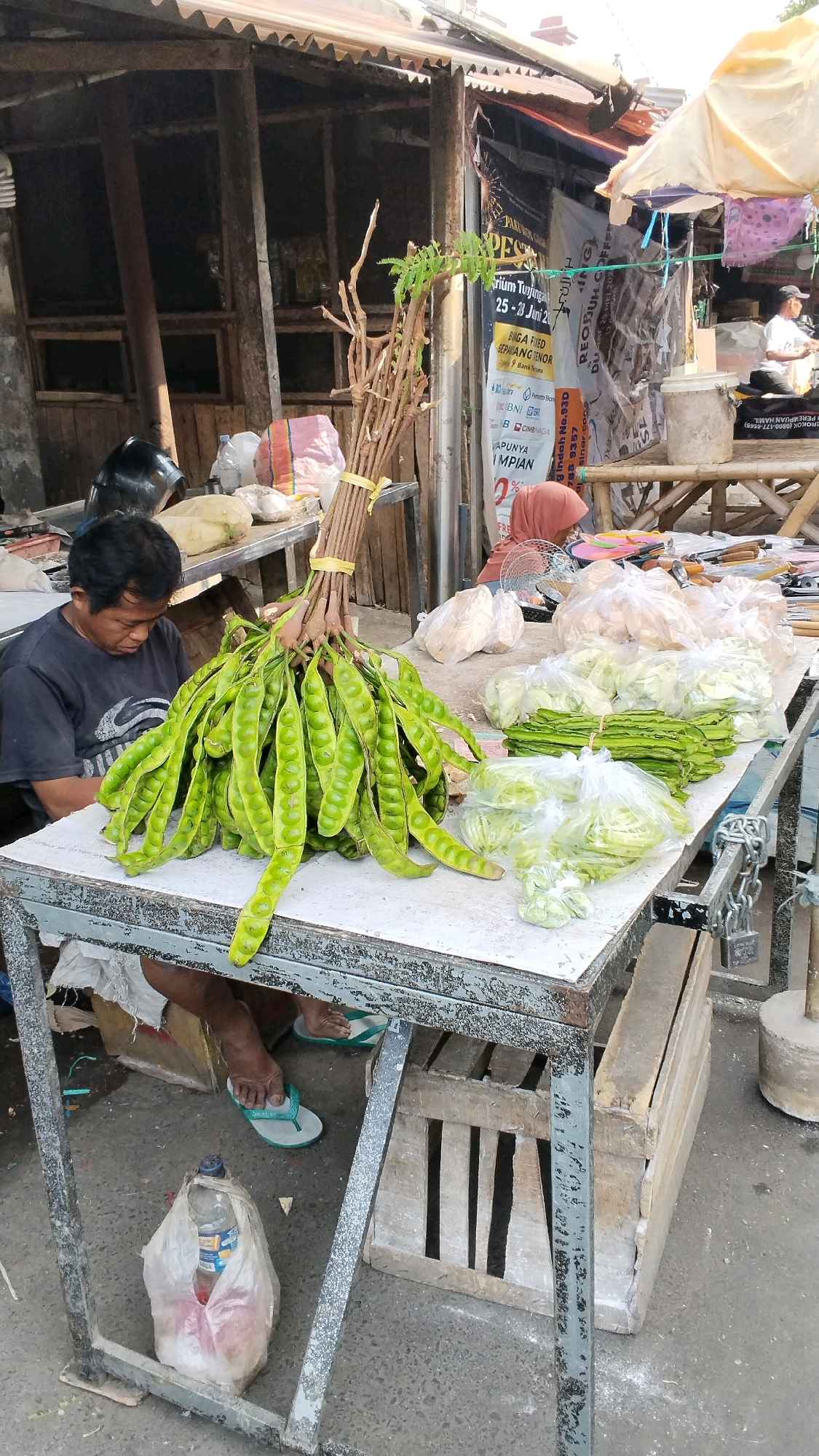 This is a full-shot, eye-level, outdoor photograph taken at a market stall in Mojokerto, Indonesia. In the foreground, a man wearing a black t-shirt and shorts sits behind a table laden with fresh produce. He is looking down at a large bunch of green petai beans, which are tied together and hang prominently in the center of the image. To the right of the petai, various other vegetables are displayed in plastic bags, including what appears to be winged beans and cabbage. Behind the man and the stall, a rustic wooden structure with a corrugated metal roof forms the background. The stall is covered by an umbrella, and various posters and banners are visible on the structure. The ground is paved and appears to be dirt and concrete. There is a plastic bag with a water bottle and other items on the ground beneath the table.