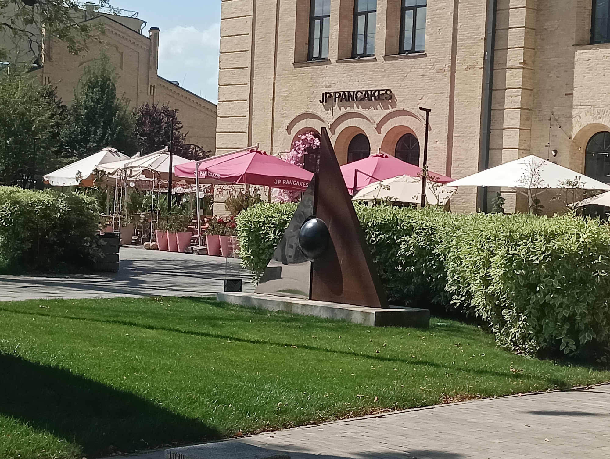The image depicts an outdoor scene in front of a brick building with the sign "JP PANCAKES" above arched windows. In the foreground, a modern abstract sculpture featuring metallic triangular shapes and a dark sphere is situated on a concrete base. Surrounding the sculpture is a manicured green lawn. Behind the sculpture, several pink and white umbrellas shade outdoor seating areas with tables and chairs, and potted plants. Dense green bushes line the area between the seating and the building. The day is sunny, casting shadows on the lawn and the paved walkway in the lower right of the frame. The overall setting appears to be a cafe or restaurant patio.