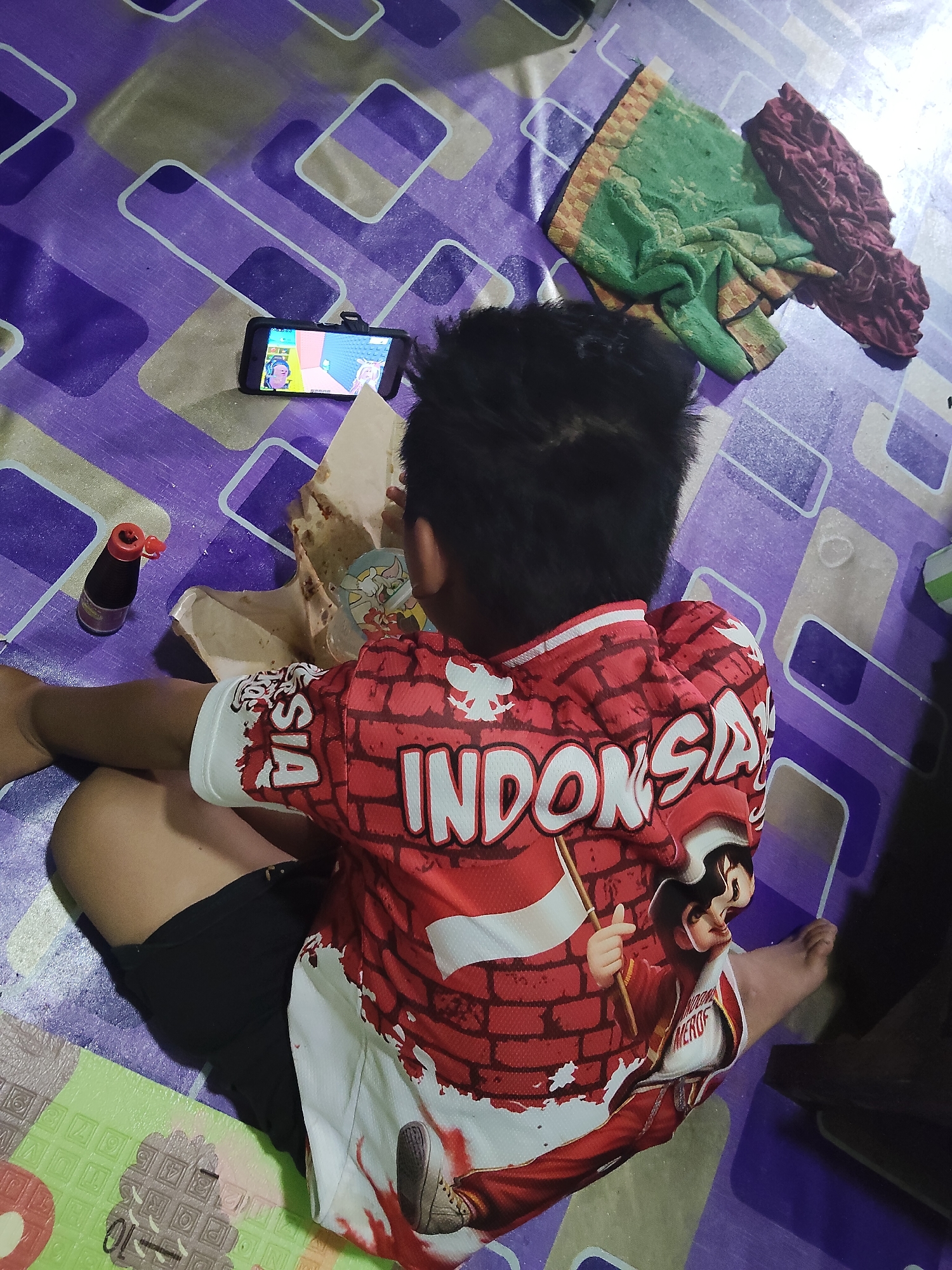 A young boy sits on a purple and grey patterned mat, facing away from the camera, engrossed in a mobile game displayed on a smartphone placed in a stand. He is wearing a red shirt with "INDONESIA" prominently displayed in white brick-like lettering. The shirt also features graphics of a person holding a flag and a person in uniform. He is wearing black shorts and his bare legs are visible. To his left, a bottle of dark liquid with a red cap sits on the mat, and next to it is a paper container of food, possibly with a strawed cup inside. To his right, two folded pieces of cloth, one green and one burgundy, are laid out on the mat. The setting appears to be indoors.