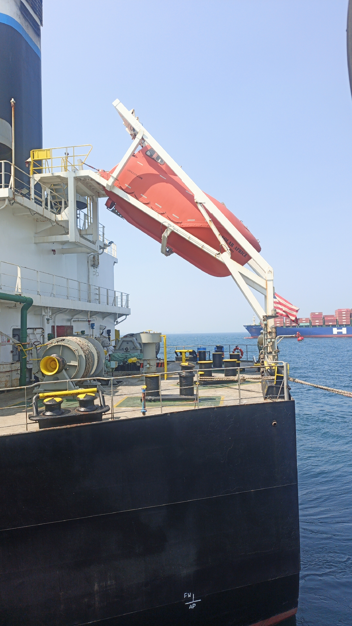 In the city of Singapore, Singapore, this image captures a view of the starboard side of a large black vessel, likely a cargo ship, at sea. A bright red lifeboat is suspended from a white davit mechanism extending from the vessel's deck. The lifeboat is angled downwards, suggesting it is either in use or prepared for deployment. In the background, a large container ship, also at sea, is visible on the horizon under a clear blue sky. The deck of the foreground vessel is equipped with various nautical equipment, including winches, ropes, and bollards. The water is calm and blue. The overall scene depicts a maritime environment.