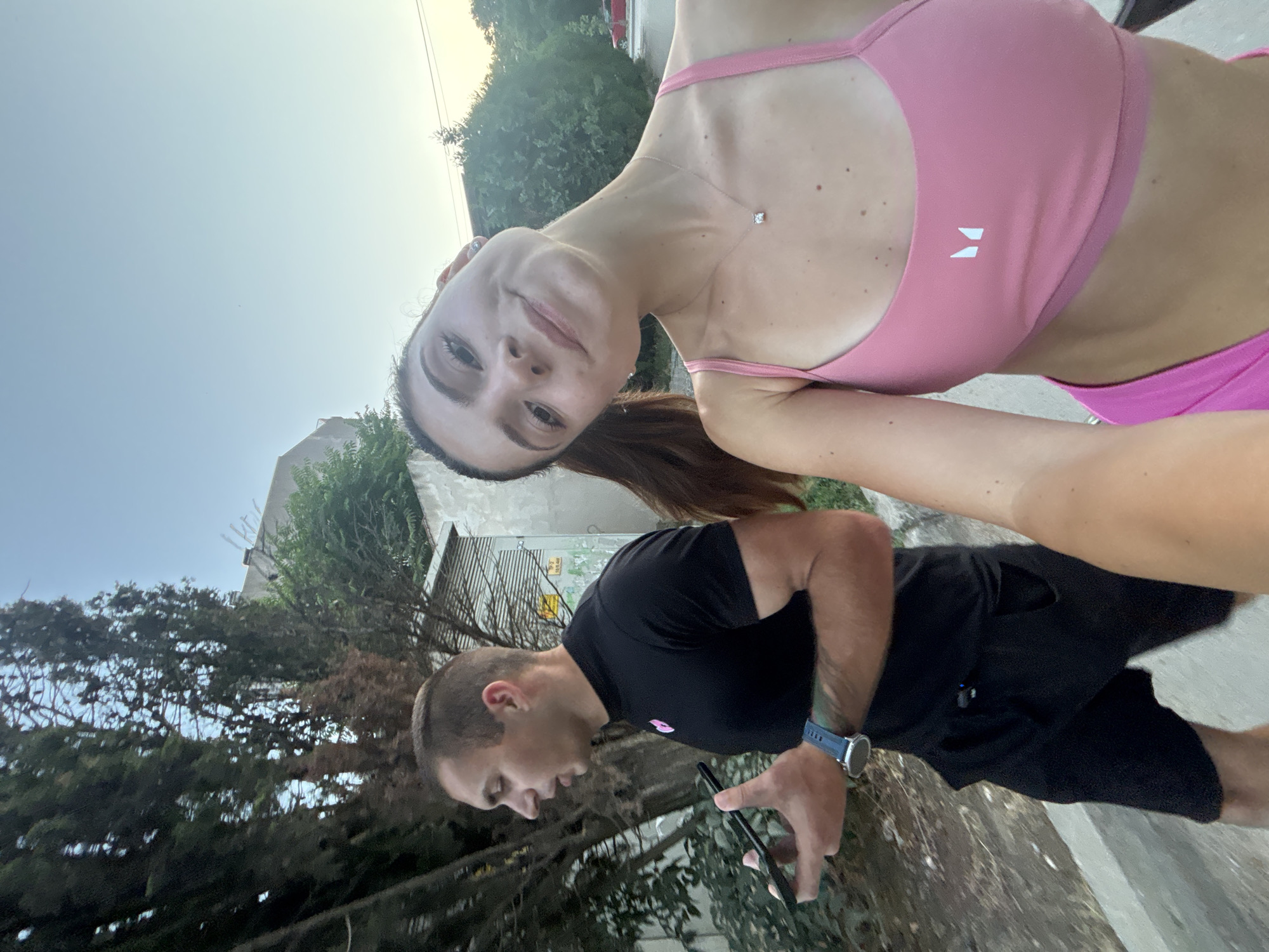 In the city of Varna, Bulgaria, on an outdoor paved walkway adjacent to a street with parked cars and trees, a young couple is captured in a selfie. The woman, in the foreground, wears a pink sports bra and matching shorts, a delicate necklace adorning her neck. The man, behind her and slightly to the left, is dressed in a black t-shirt and shorts. He holds a mobile phone in his left hand and a smartwatch is visible on his left wrist. The background reveals a building and dense foliage. The sky is clear and bright, suggesting daytime.