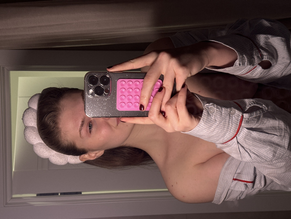 A female subject is depicted taking a mirror selfie indoors in Бакча-Сарай, Russia. The individual, a young adult female, has the right side of her face visible, characterized by a faint smile and a white, textured, bubble-style headband holding back her hair. Her left face is obscured by a smartphone, held in both hands. The phone, which appears silver-toned, is enclosed in a clear, glitter-infused case and features a pink silicone suction-cup array on its reverse. Her fingernails are painted dark red. She is dressed in a light-striped, off-the-shoulder shirt with red trim detailing on the cuffs, exposing her bare shoulders and upper torso. A repeating circular pattern is faintly visible on her upper chest. The immediate background shows a light-colored doorframe to the left and a dark fabric curtain to the right. The scene is dimly lit, with portions in shadow.
