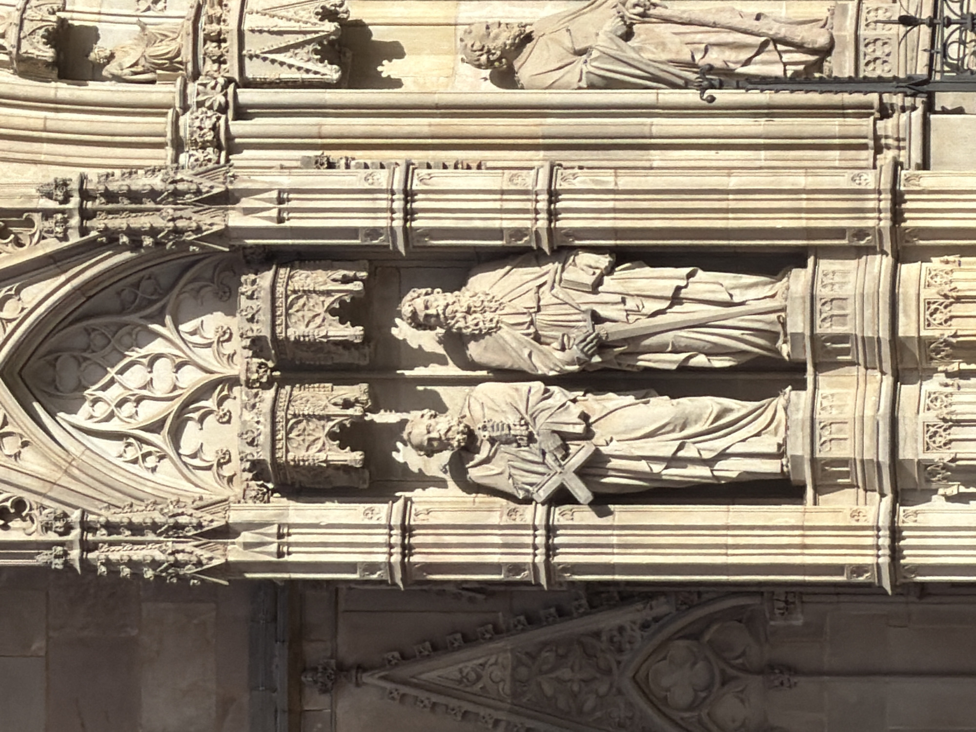 This image shows a detail of the facade of the Barcelona Cathedral, located in Barcelona, Spain. The photograph focuses on a section of Gothic architecture featuring two large stone statues of bearded men in flowing robes. Each statue stands on a decorative pedestal. The statue on the left holds a cross-shaped object in its hands. The statue on the right holds a scroll or book. The statues are positioned within niches and are flanked by fluted columns and ornate architectural embellishments, indicative of Gothic design. Sunlight casts shadows on the stone, highlighting the intricate details of the sculptures and the building's facade.