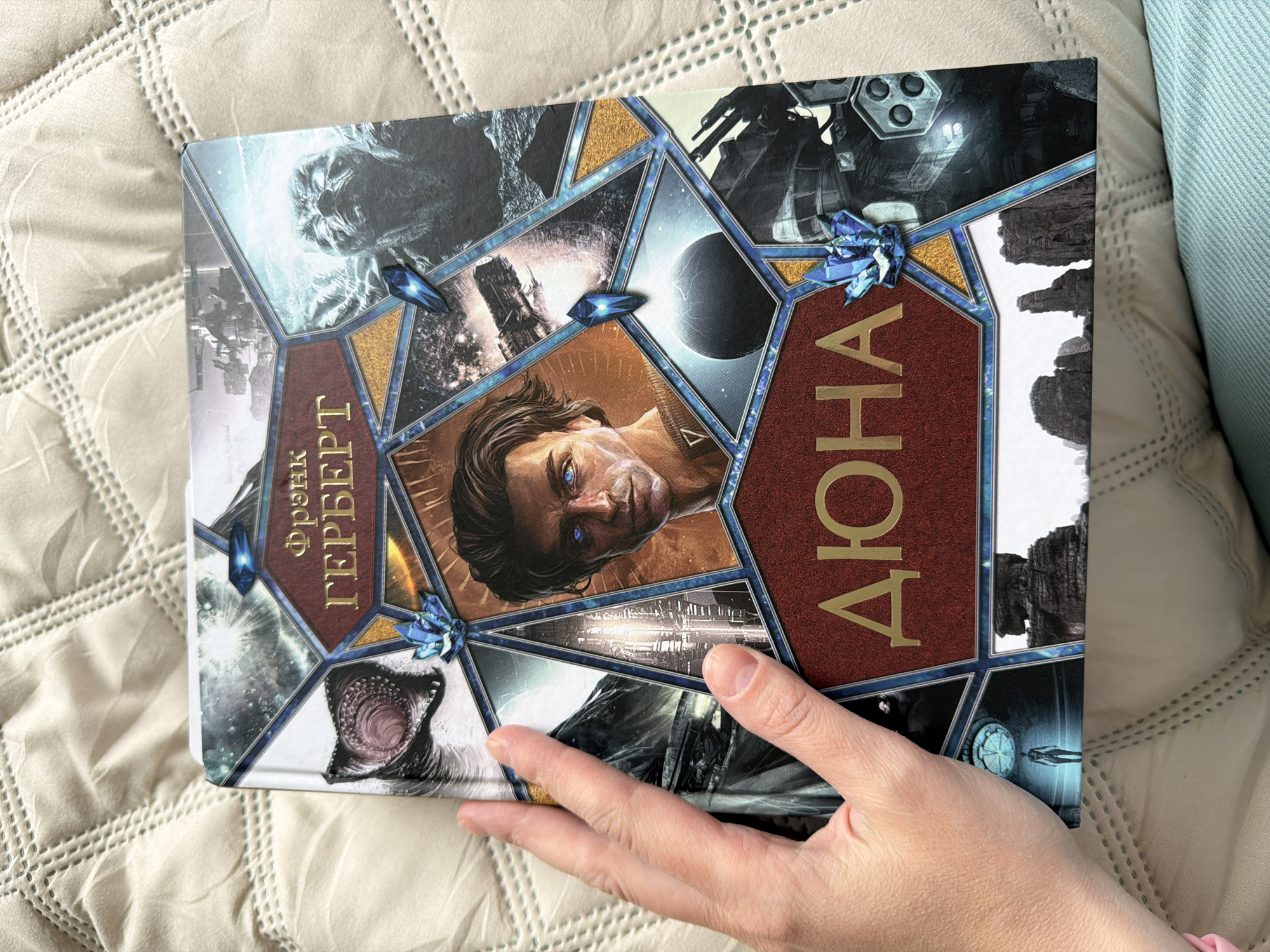 A person's hand holds a copy of the Russian-language edition of Frank Herbert's "Dune" book. The book cover features a mosaic-style design with images related to the Dune universe, including Paul Atreides, a sandworm, spaceships, and desert landscapes. The book is lying on a quilted beige fabric with a slight shimmer. In the upper right corner, a light blue fabric is also visible.
