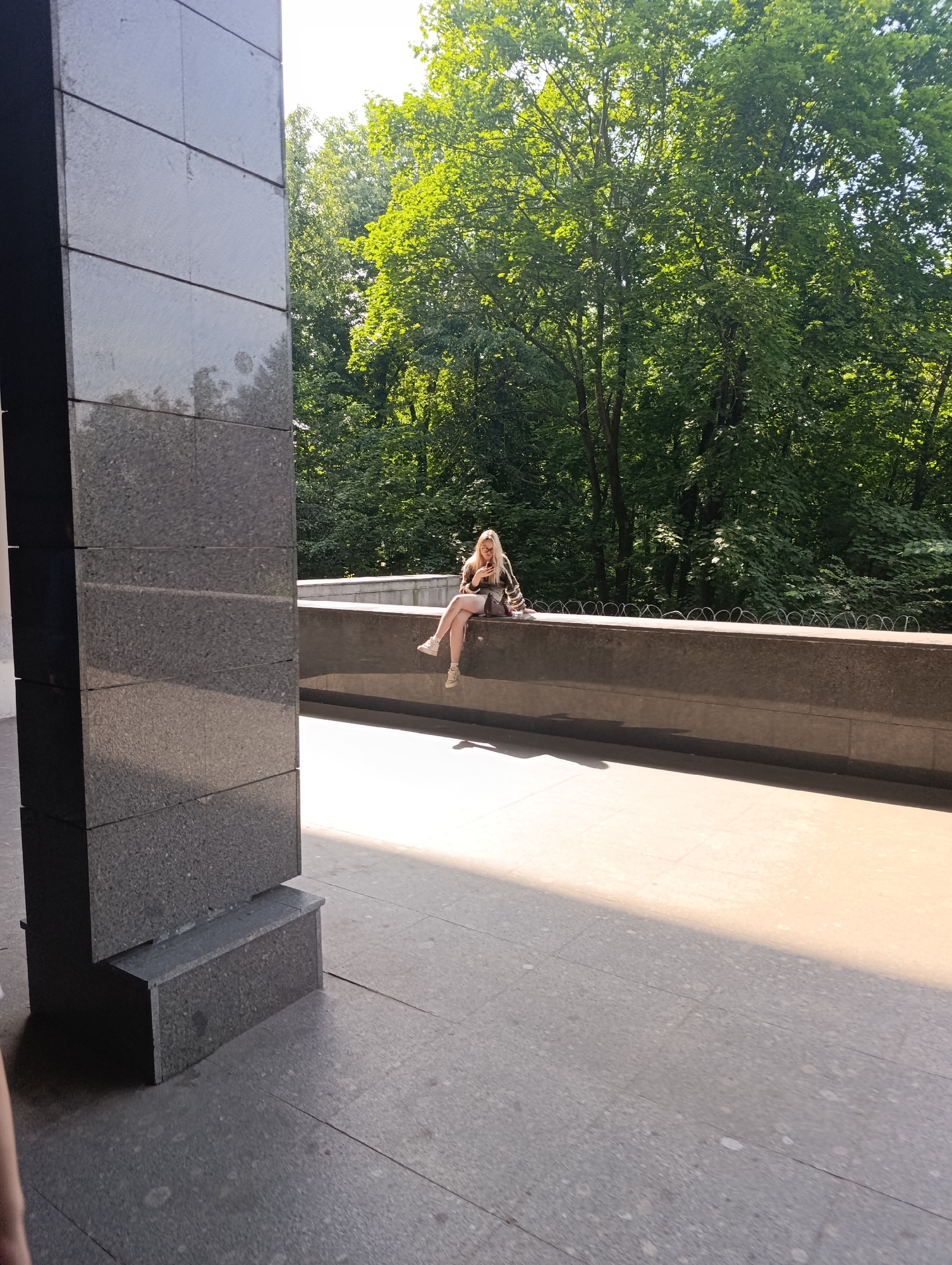 The image depicts an outdoor scene on a sunny day. A blonde woman with glasses is seated on a dark grey, stone barrier or wall that extends horizontally across the mid-ground. She is wearing a light-colored, patterned long-sleeved top and dark shorts, with light-colored sneakers on her feet. She is holding a smartphone in both hands and looking down at its screen. Behind her, a dense expanse of vibrant green trees, characteristic of a forest or park, fills the background. On top of the stone barrier, behind the woman, a line of coiled razor wire is visible. To the left, a large, dark grey architectural column, composed of smooth, rectangular stone blocks, occupies a significant portion of the foreground and mid-ground. A smaller, step-like structure of similar material is at the base of this column. The ground in the foreground is paved with light grey, speckled stone tiles. A sharp diagonal shadow from an unseen overhead structure or object cuts across the tiled ground, with the area to the right of the shadow bathed in bright sunlight.