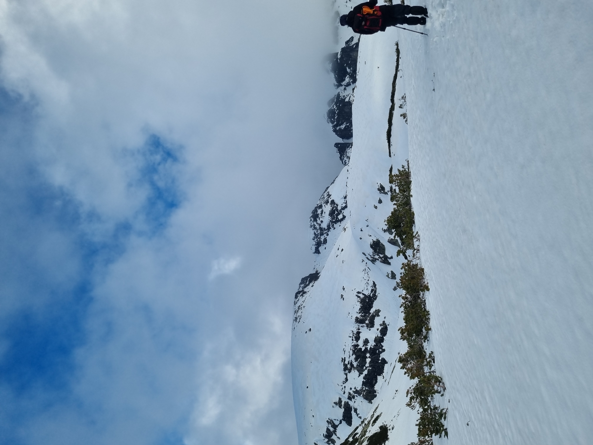 The image depicts a snowy mountain landscape near Govedartsi, Bulgaria. The sky is partly cloudy. A person wearing dark clothing and a backpack with orange accents is standing on a snow-covered slope, holding a walking stick. The mountain has patches of bare rock and scattered trees. The snow-covered slope extends to the right side of the image.