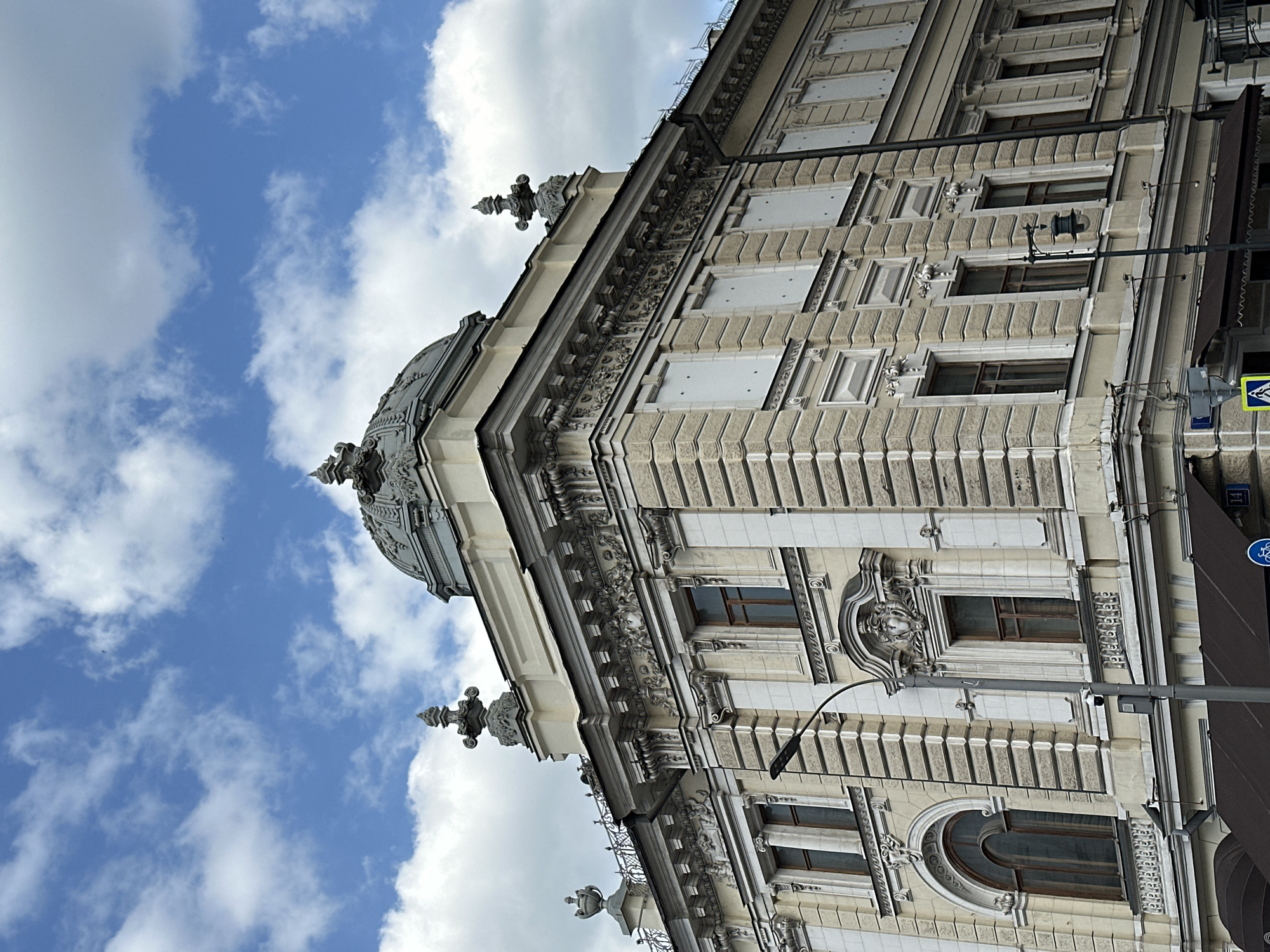 The image captures the architecture of a building in Moscow, Russia. It showcases the corner of a structure with a dome, embellished with ornate detailing and sculptures. The building's facade features textured, layered walls and windows, alongside light fixtures and a sign indicating a pedestrian crossing. The sky is visible, displaying a clear blue backdrop with scattered, puffy white clouds.