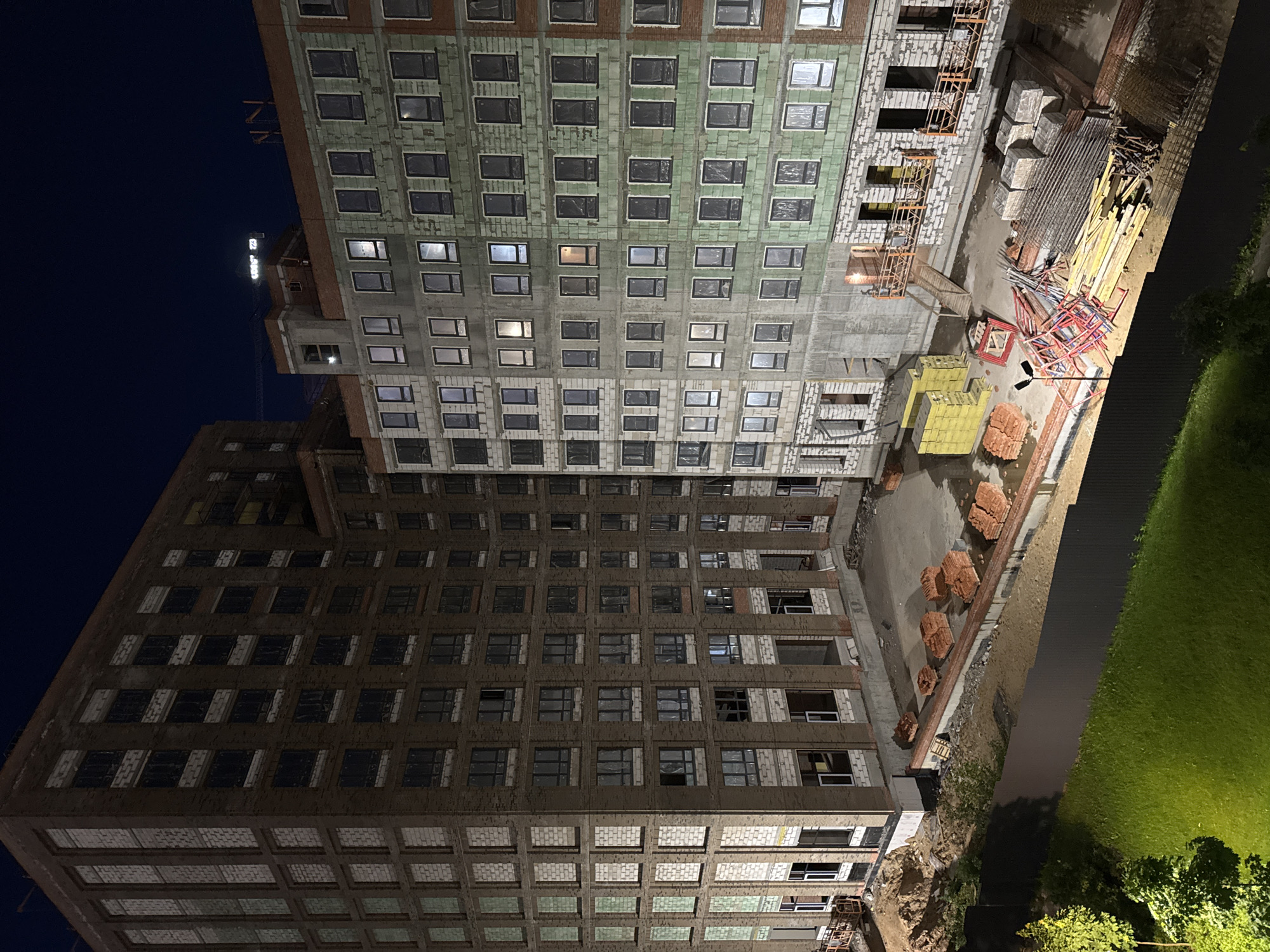This nighttime photograph, taken in Izhevsk, Russia, depicts an active construction site featuring two large, multi-story residential buildings in different stages of completion. The building on the left appears darker, with exposed concrete and brickwork, and a grid of installed windows. The building on the right is more illuminated, showcasing a facade with partially completed sections, numerous windows (some with lights on inside), and scaffolding attached.
Between and around the buildings, the ground is scattered with various construction materials, including stacks of red bricks, bundles of lumber, metal rods, yellow insulation panels, and concrete blocks. Red and yellow metal scaffolding structures are also visible on the ground. A bright yellow crane arm extends into the dark night sky in the background, hinting at ongoing heavy construction. A street lamp provides illumination to part of the construction area. On the far right, a strip of brightly lit green grass and some trees stand out against the darkness.
The time of day is night, indicated by the deep blue-black sky and the reliance on artificial lighting from the buildings' windows, the street lamp, and construction site equipment. The weather appears clear. No people are visible in the image, and no discernible text can be read on any of the structures or materials. The scene captures an urban development project under construction.