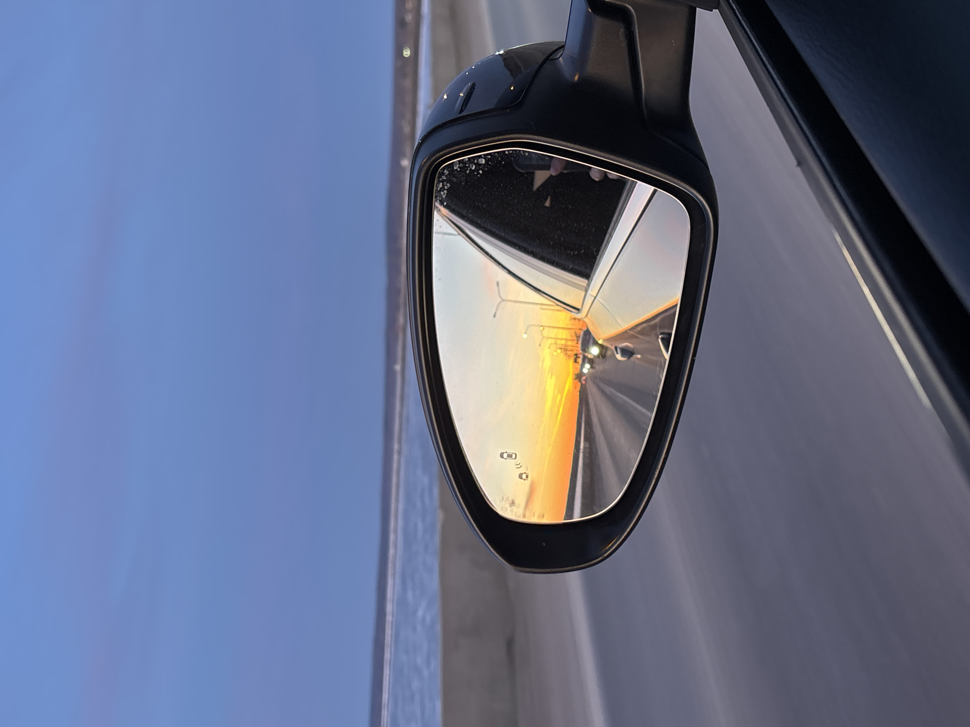 A close-up, horizontally oriented shot captures the side mirror of a moving car, offering a dynamic view of a highway at sunset. The dominant feature is the black, angular side mirror, positioned against a clear, pale blue sky on the left and the dark, slightly blurred body of the car and road on the right, suggesting motion.
Inside the mirror's reflection, a brilliant sunset bathes the scene in vibrant orange and yellow hues, casting a warm glow over the reflected road ahead. Streaking lines on the road surface, visible within the reflection, further emphasize the vehicle's speed. Several bright streetlights are also reflected along the highway, hinting at an urban or suburban setting.
In the lower-left corner of the mirror's reflective surface, two distinct white icons are visible, representing a blind spot monitoring system. These icons, depicting small car shapes with signal lines, indicate active safety features. The mirror's housing and outer edges show minor speckling, possibly from dust or water spray. The overall impression is one of travel during a picturesque dusk, with advanced vehicle technology highlighted. The location is Chaikovsky, Russia.