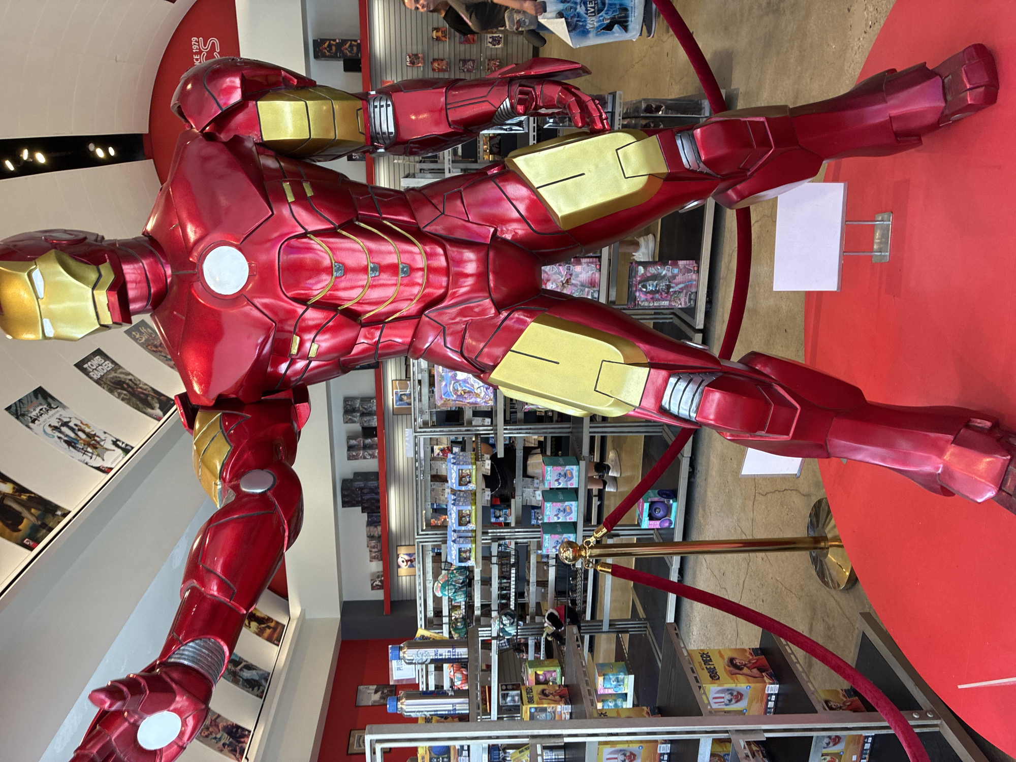 An Iron Man statue, approximately 7-8 feet tall, is displayed prominently on a red circular platform in a store. The statue is in its iconic red and gold armor, with its left arm extended forward and its right arm at its side. Behind the statue, shelves are stocked with various merchandise, including toys, action figures, and collectibles. A red velvet rope barrier guides visitors around the statue. In the background, two women are visible, one browsing the shelves and the other carrying a shopping bag. The store appears to be a retail establishment specializing in pop culture and collectible items, possibly a comic book or toy store.