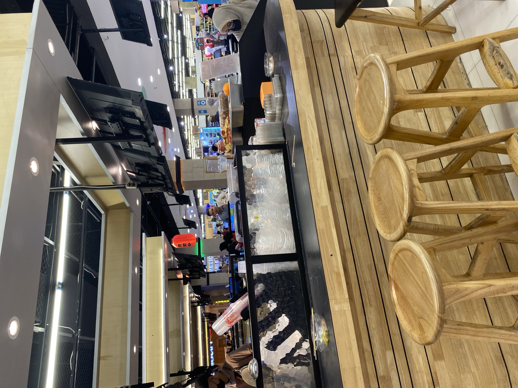 The image depicts the interior of a food establishment, possibly a cafe or restaurant, within a commercial setting. In the foreground, three wooden bar stools are visible, two of which are fully in view. Behind them, a counter is constructed from light-colored wood with a dark countertop. On the countertop, stacked plastic cups, small containers of food, and chopsticks are arranged. A person wearing a black mask and an apron is standing behind the counter, partially visible. In the background, several individuals are interacting, and shelves stocked with goods are lined up. Suspended from the ceiling are multiple flat-screen televisions. A red Japanese lantern hangs from the ceiling. The overall environment suggests a well-lit, modern retail space. The presence of specific items like the Japanese lantern and the food presentation may indicate a Japanese-themed eatery. The scene is observed from a slightly elevated angle, looking across the counter and into the wider space.