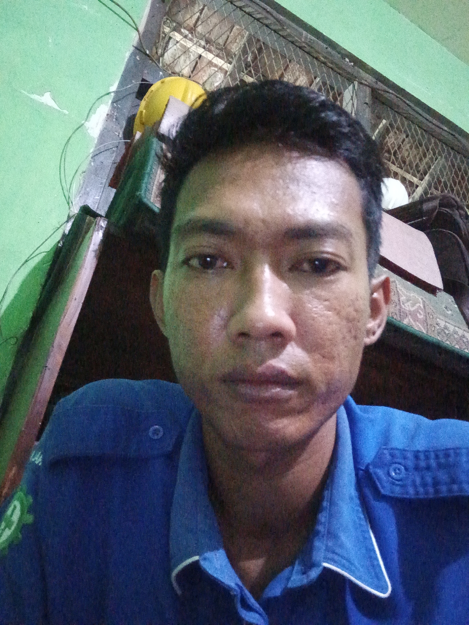 The image is a selfie of a young man wearing a blue collared shirt with a button and pocket on the chest. He has dark hair and appears to be in an indoor setting. The background shows a green wall, a metal grate, and some objects that are difficult to identify clearly due to low resolution and lighting. There is a yellow hard hat visible on a shelf or surface behind him, along with what appears to be a patterned textile. The man's face is the primary focus, and his skin has visible texture and some imperfections. The overall impression is of an informal, personal photograph taken in a somewhat cluttered environment, possibly a workshop or a storage area. The location in Kidul Dalem, Indonesia, is not discernible from the visual information in the image itself.