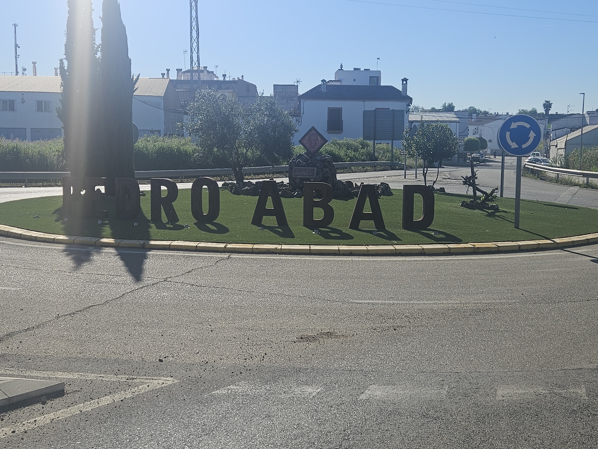 The image captures a sunny day scene in Pedro Abad, Spain. At the foreground is a roundabout with the large metal letters spelling "PEDRO ABAD" displayed on the green area in the center. There is a sign indicating that the area is a roundabout. Behind this, the town’s architecture is visible, with houses and buildings under a clear sky. The road leading up to the roundabout shows typical signs of use. A couple of tall, thin trees partially obscure the "PEDRO" portion of the name. A small monument is located behind the letters.