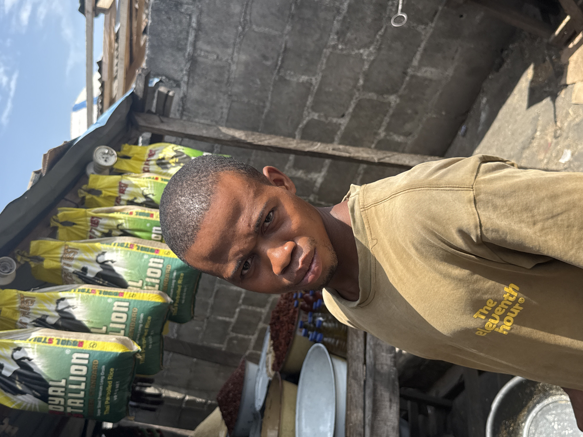 A close-up photo shows a man in front of a stall with bags of rice. The man is wearing a brown t-shirt. On the t-shirt, the words "The eleventh hour" are printed. Behind him, stacked vertically, are bags of "Royal Stallion" rice. The bags are primarily green and yellow with a picture of a horse on them. Next to the rice, there are various bowls and containers. The background shows a partially constructed or unfinished building made of concrete blocks. The sky is visible in the upper left corner of the image. The image appears to be taken at the city of Ado, Nigeria.