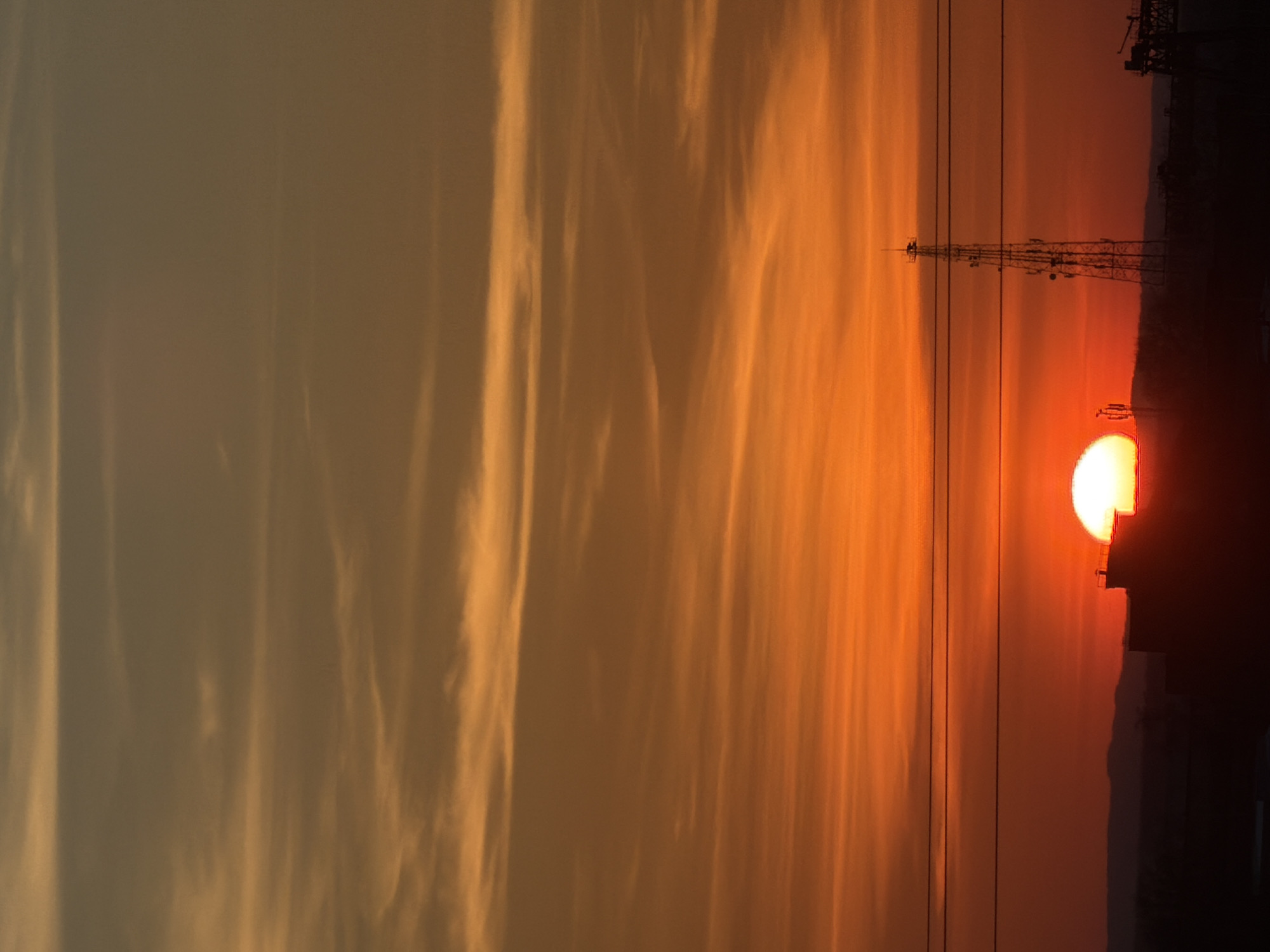 The image depicts a sunset in an urban area, likely in the vicinity of Chaikovsky, Russia. A partially obscured sun, a power transmission tower, overhead power lines, and the silhouette of buildings against a fiery orange and golden sky are visible. Streaks of clouds are visible to the left. The time of day is late afternoon or early evening. There are no discernible people or interactions in the scene.