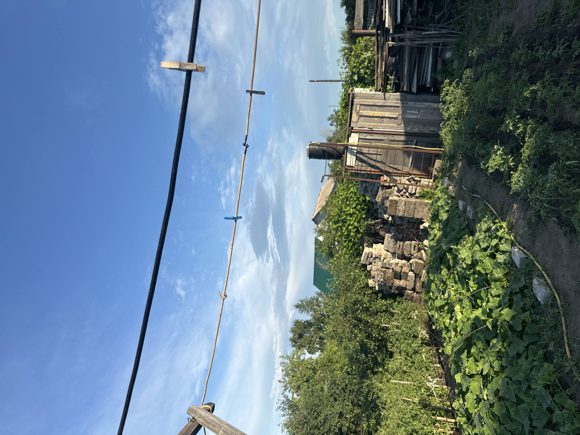 This is a landscape photograph taken outdoors on a sunny day. The image is oriented vertically. In the foreground, a black cable runs diagonally from the bottom left towards the top right. Attached to a thinner clothesline suspended below the cable are several clothespins, some wooden and some blue. The background features a rural scene. To the right of the cable, there's a weathered wooden structure, possibly a shed or outbuilding, with a black water tank on top. Piles of bricks and building materials are scattered around the base of this structure. Beyond that, lush green vegetation, including trees and what appears to be a garden patch with climbing plants, fills the right side of the frame. In the distance, the sky is a vibrant blue with scattered white clouds. The overall impression is one of a rustic, possibly agricultural setting, bathed in bright sunlight, suggesting it is daytime. The location is identified as Tobyl, Kazakhstan. There is no visible text in the image.