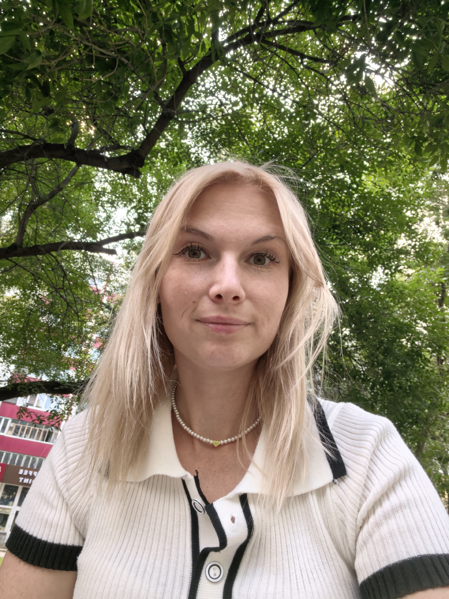 In this frontal medium shot, a young Caucasian woman with blonde hair and light skin poses for a selfie outdoors. She is wearing a white collared shirt with dark green trim on the collar and sleeves, and a pearl necklace with a small green heart pendant. She has light brown eyes and a neutral expression. Behind her, lush green tree branches create a canopy, with glimpses of a building with red and white accents visible through the leaves. The image appears to be taken in an urban park or courtyard in Sterlitamak, Russia.