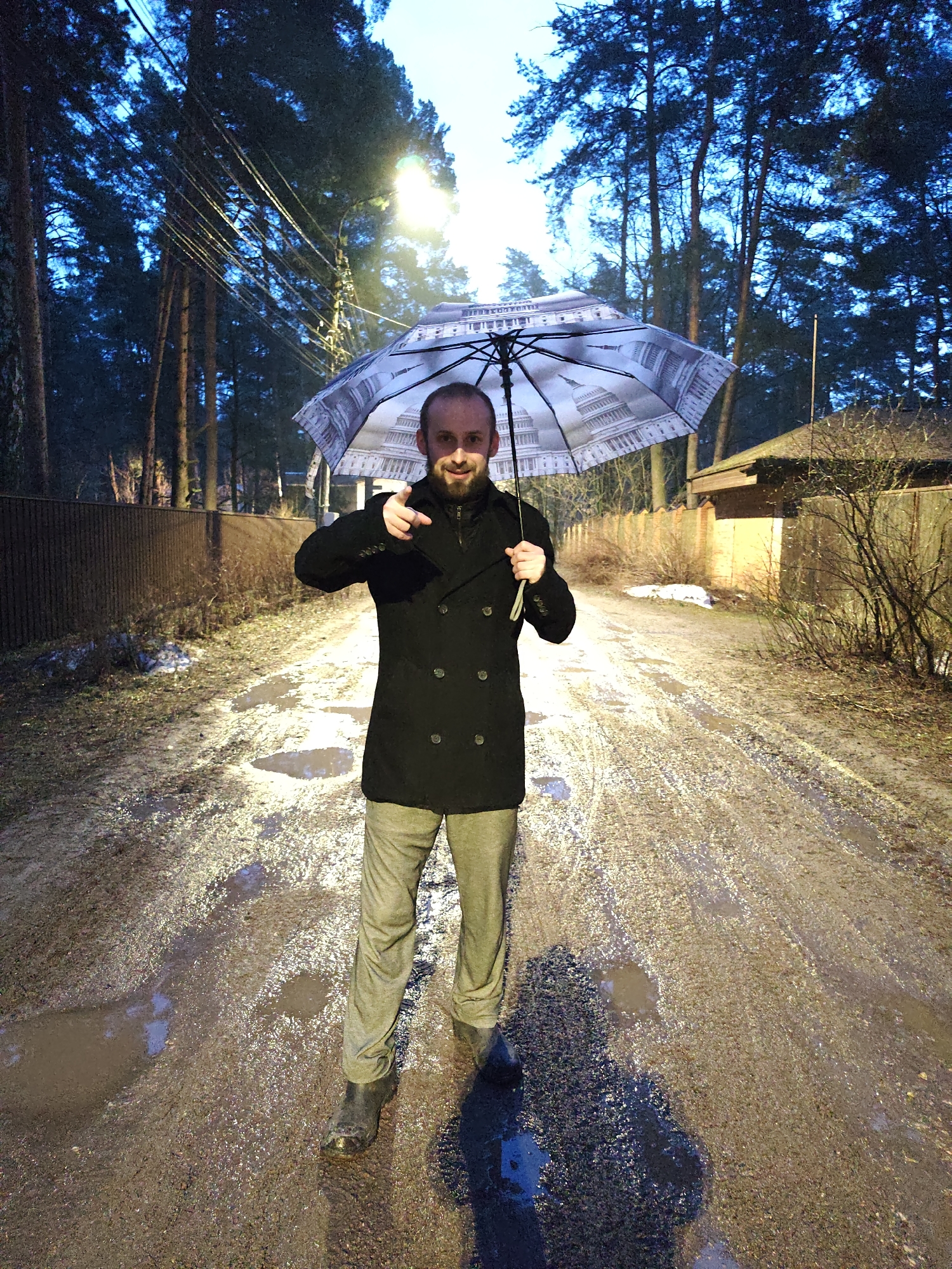 A bearded man stands on a muddy, puddle-filled road under the light of a street lamp in what appears to be a residential area. The man is holding an umbrella decorated with an image of the United States Capitol building, and he is pointing towards the viewer with his right hand. He is wearing a dark double-breasted coat, light-colored trousers, and dark boots. The background shows a fence, buildings, and tall trees. The overall lighting suggests it is either late evening or early morning. The city is Kratovo, Russia.