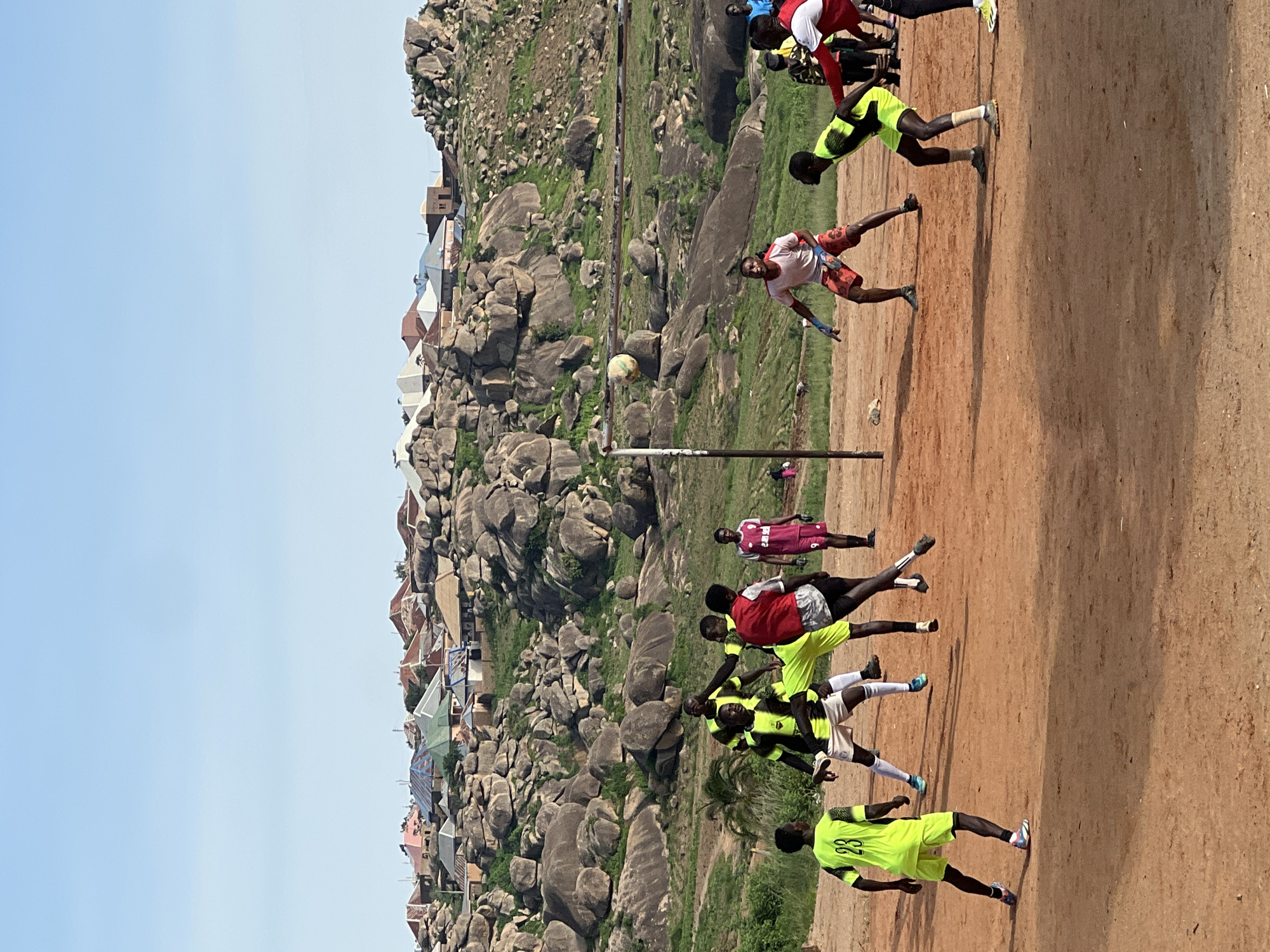 The image shows a group of young men playing soccer on a dirt field in Jos, Nigeria. The field is located in a rocky, hilly area with houses visible in the background. The sky is clear and blue. The players are wearing athletic attire, and some are in mid-action, kicking or jumping for the ball. The game appears to be a casual match, with no formal equipment like goalposts or nets, save for a makeshift net in the center. The atmosphere seems energetic and playful, with the players focused on the game. The rocky landscape of Jos is a prominent feature of the background, with a cluster of buildings nestled among the rocks.