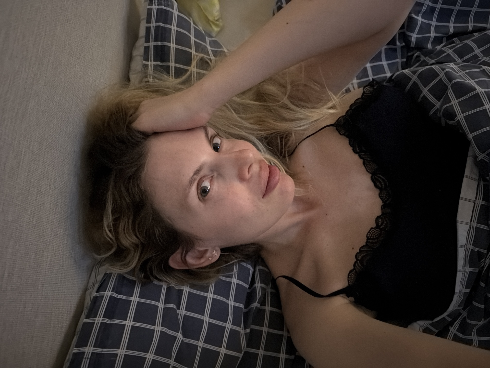 A young adult female is lying in bed. She is positioned on her left side, facing the camera with her head tilted. Her left arm is raised, with her hand resting on her forehead. The woman has fair skin, blonde hair, and is wearing a black camisole with lace trim. She has an earring visible in her left ear. The bed has a blue and white checkered pattern. The scene appears to be indoors, likely a bedroom. The image was not taken in Budva, Montenegro.