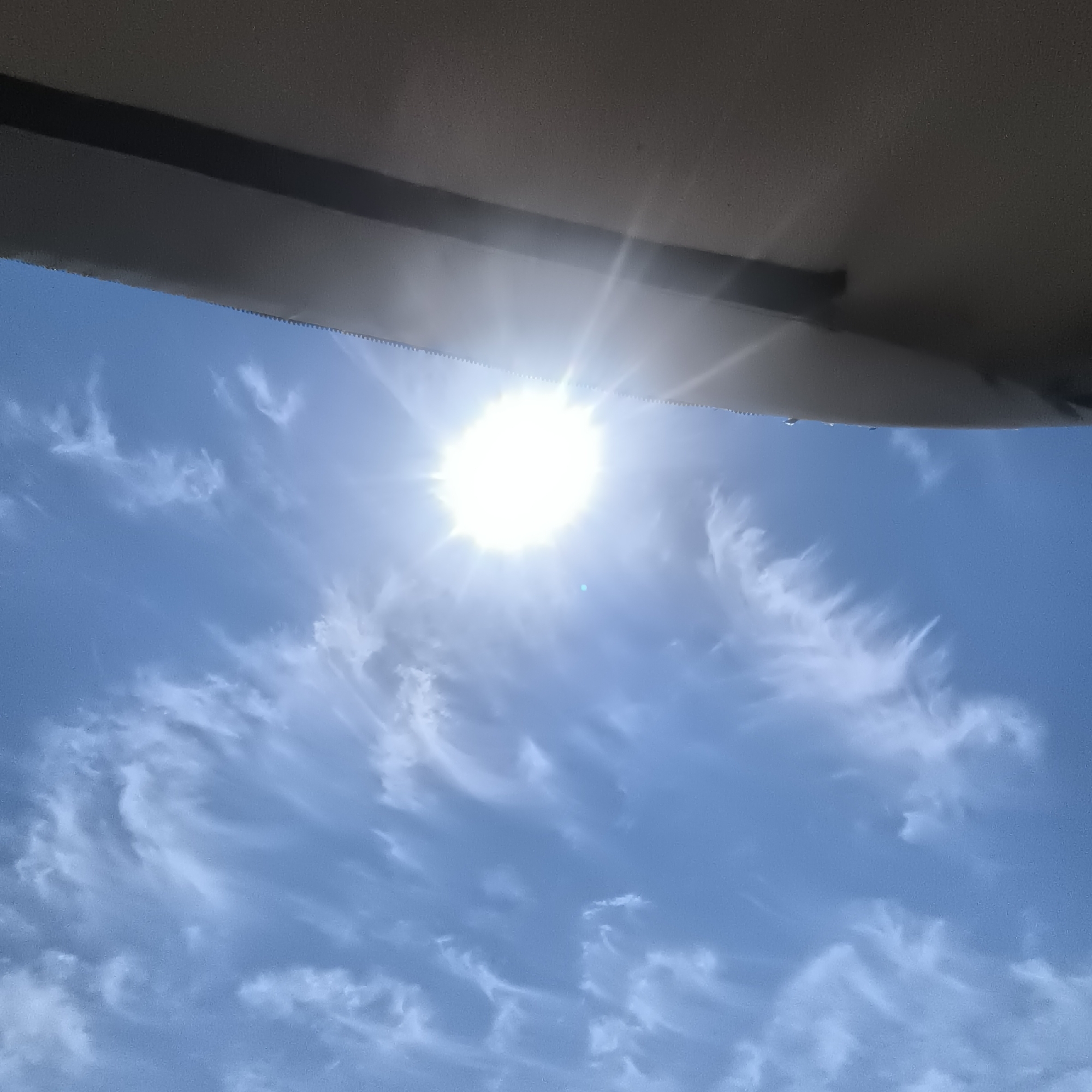 This is a low-angle, medium shot of a bright sun in a clear blue sky with scattered wispy white clouds. A dark, textured surface with a serrated edge is visible at the top of the frame, obscuring a portion of the sky. This object appears to be the interior of a vehicle's roof or a similar structure. No people are visible in the image. The scene suggests a sunny day observed from within a vehicle. The provided location of Malyy Baklaniy, Russia, is not visually determinable from the image itself.