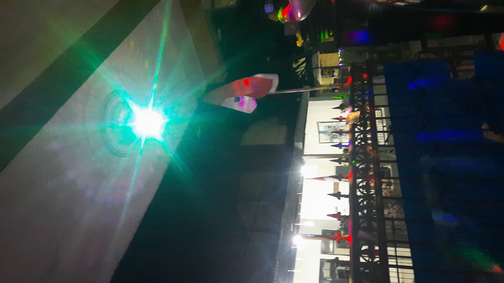 The image shows a flag, possibly the Indonesian flag, waving in front of a building at night. There is a bright green light shining down from above, creating a starburst effect. The building appears to have lights on inside and is fenced off with decorative metalwork. Some small flags or decorative lights are visible along the fence. The overall scene suggests an outdoor event or gathering. The location is Sidoarjo, Indonesia.