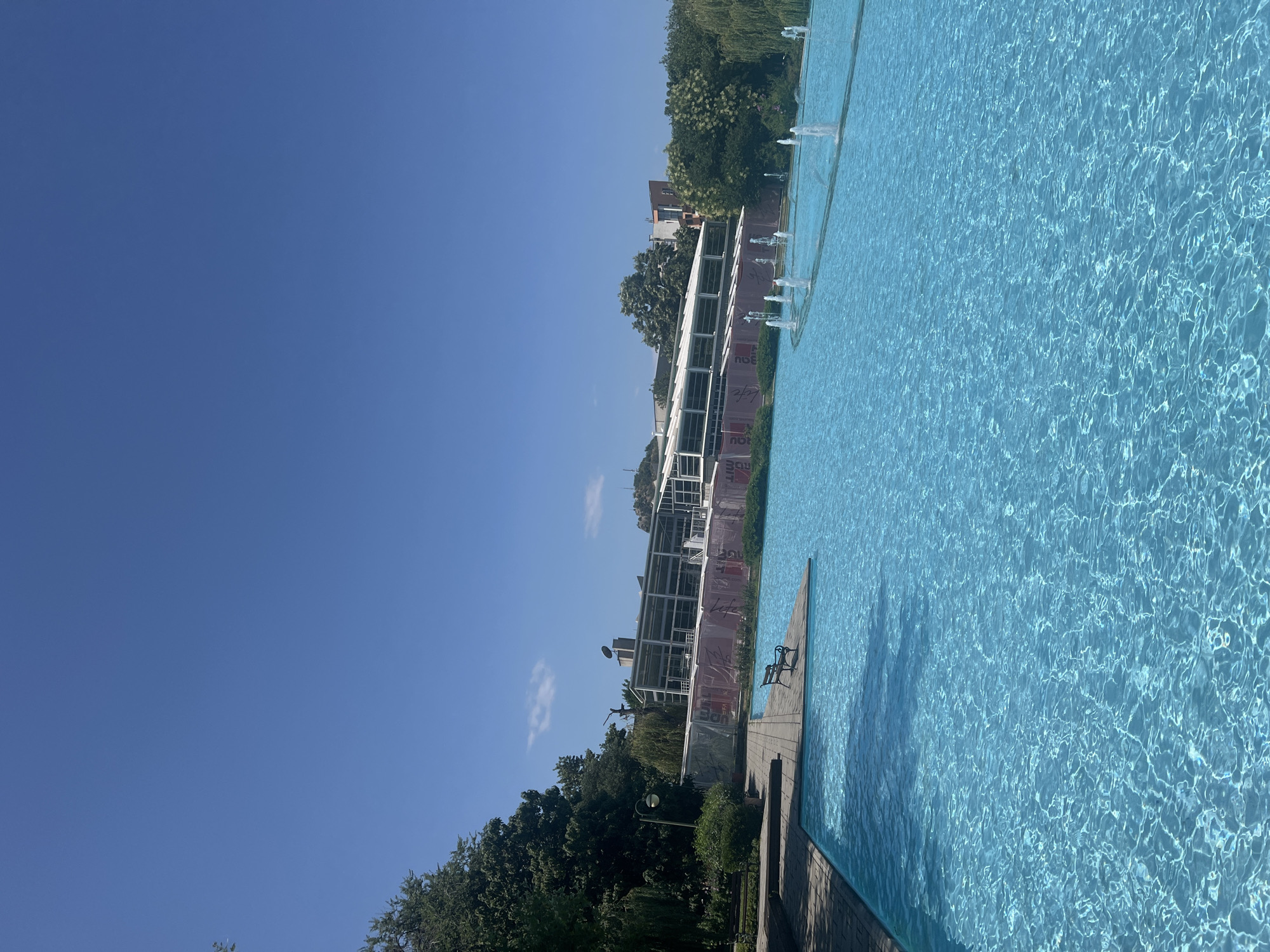 The image depicts a large, bright blue swimming pool or decorative fountain area on a sunny day. The water is clear and shows ripples from numerous small water jets spraying upwards in a curved line across the expanse. In the foreground, a paved walkway with a wooden deck juts out into the water, featuring a single dark-colored park bench. To the left, a dense green tree provides shade over another paved area with a lamp post and some greenery.
In the background, a long, modern building with a white, sloped roof and numerous windows is visible. Red and white banners are draped across the lower section of this building, suggesting it might be a venue or exhibition space. More trees and some distant buildings, including a rocky incline with a structure on top, are visible beyond the main building. The sky is a clear, bright blue with a few wispy clouds.
This scene is located at the Singing Fountains in Plovdiv, Bulgaria, situated near the Tzar Simeon's Garden.