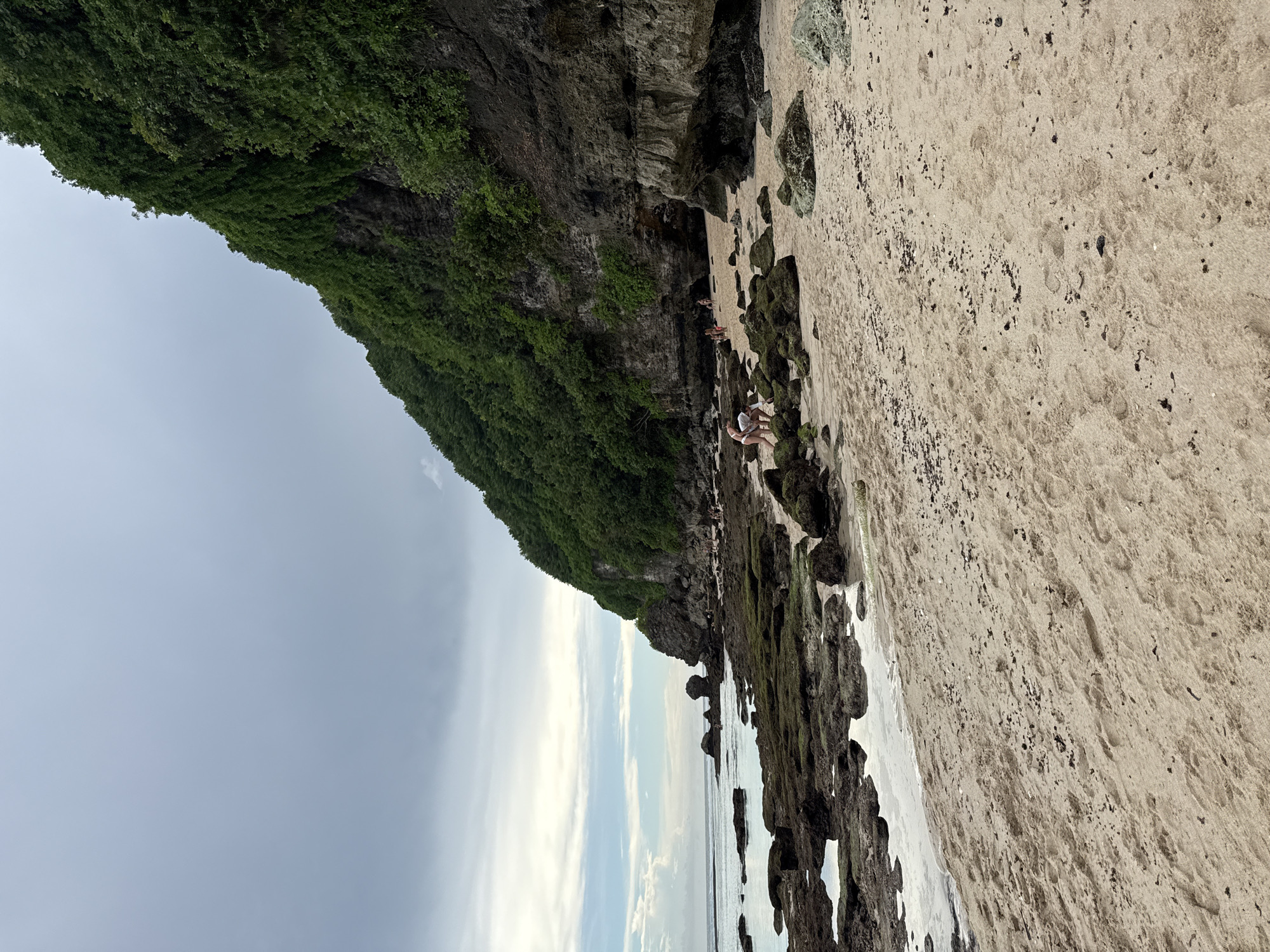 A medium shot depicts a beach at low tide located in Ungasan, Indonesia. A sandy beach stretches to the right, interspersed with dark, moss-covered rocks and tidal pools near the water's edge. The ocean and sky are visible in the left half, showing a mix of cloudy, overcast conditions with hints of blue and white along the horizon.
A cliff covered in dense green vegetation rises along the top edge of the image, forming a natural barrier between the beach and the land. Several people are congregated in the shade near the base of the cliff. One individual in the foreground is wearing light-colored clothing and appears to be crouching or kneeling. Further back, there are a few others standing or sitting in a more shaded area, closer to the rock face.