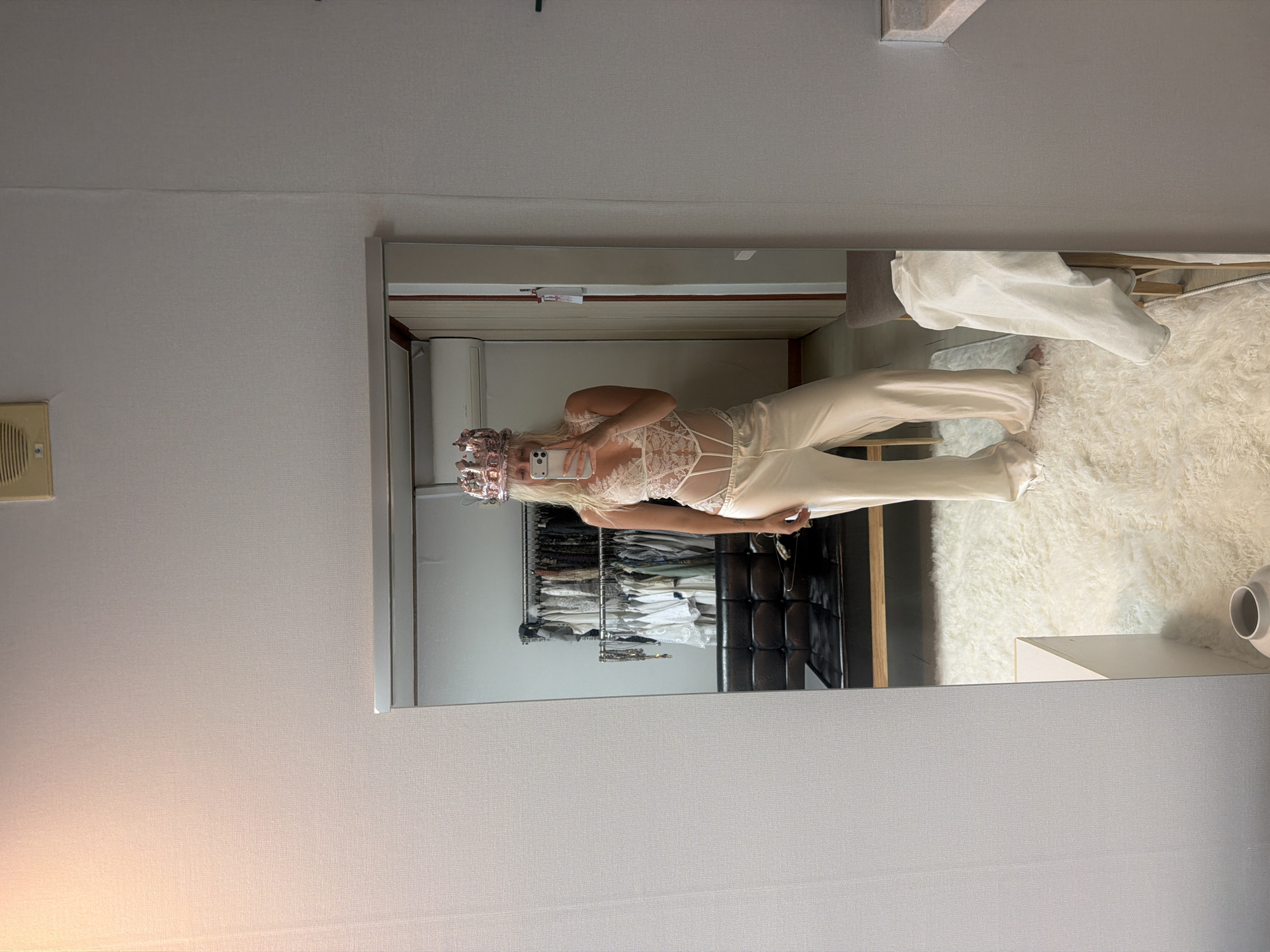 A woman is captured in a full-length mirror selfie, standing in what appears to be a bedroom or dressing area. She has long blonde hair styled with numerous large, metallic pink/rose gold hair rollers or curlers. She is wearing a cream or white lace bodysuit or top with intricate patterns and thin straps, paired with loose-fitting, cream-colored satin or silk pants. She holds a white smartphone with her right hand, taking the picture, and her left hand is casually by her side.
The mirror reflects a room with light-colored walls. Behind the woman's reflection, there is a clothing rack heavily laden with various garments, suggesting a closet or wardrobe area. A white, shaggy, and fluffy rug covers the floor in front of the mirror. Parts of a bed, possibly covered with white bedding, are visible in the reflection to the right. To the far left, a light-colored air conditioning unit or vent is mounted on the wall. A small, light-colored table or nightstand with a white cylindrical object is also partially visible on the right side of the reflection. The overall scene suggests an intimate moment of personal grooming or preparing for an event. No discernible text is visible in the image.