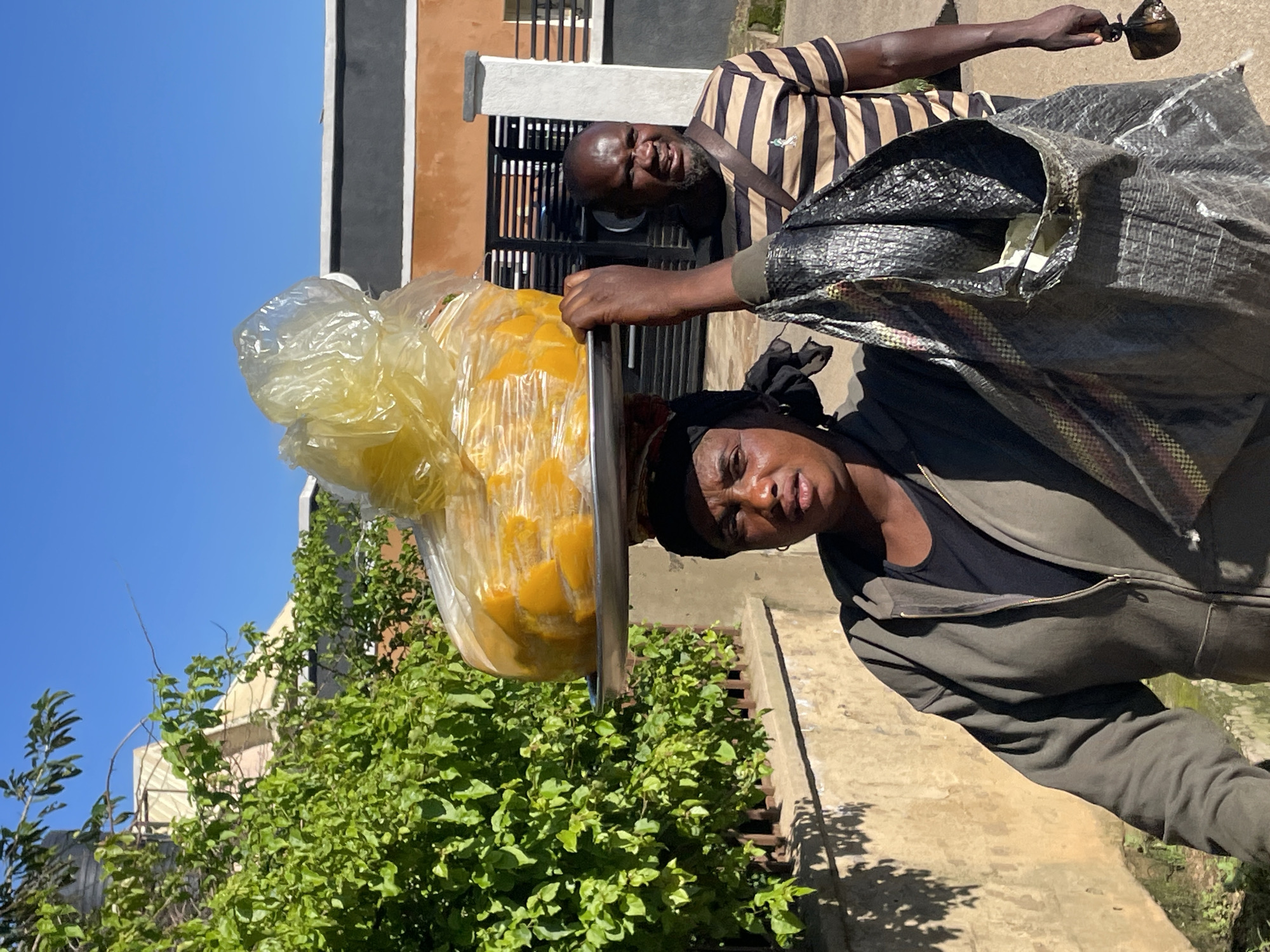 In the city of Jos, Nigeria, a woman with a headscarf walks forward, carrying a large tray of yellow produce, likely mangoes, covered in plastic wrap, balanced on her head. She wears a dark hooded jacket over a black top and holds a large, woven bag in her left arm. Behind her, a man with a striped t-shirt and a satchel walks, looking towards the camera with a tense expression. The environment appears to be an urban street with a concrete wall and lush green foliage bordering the path. The sun is bright, casting shadows on the ground.