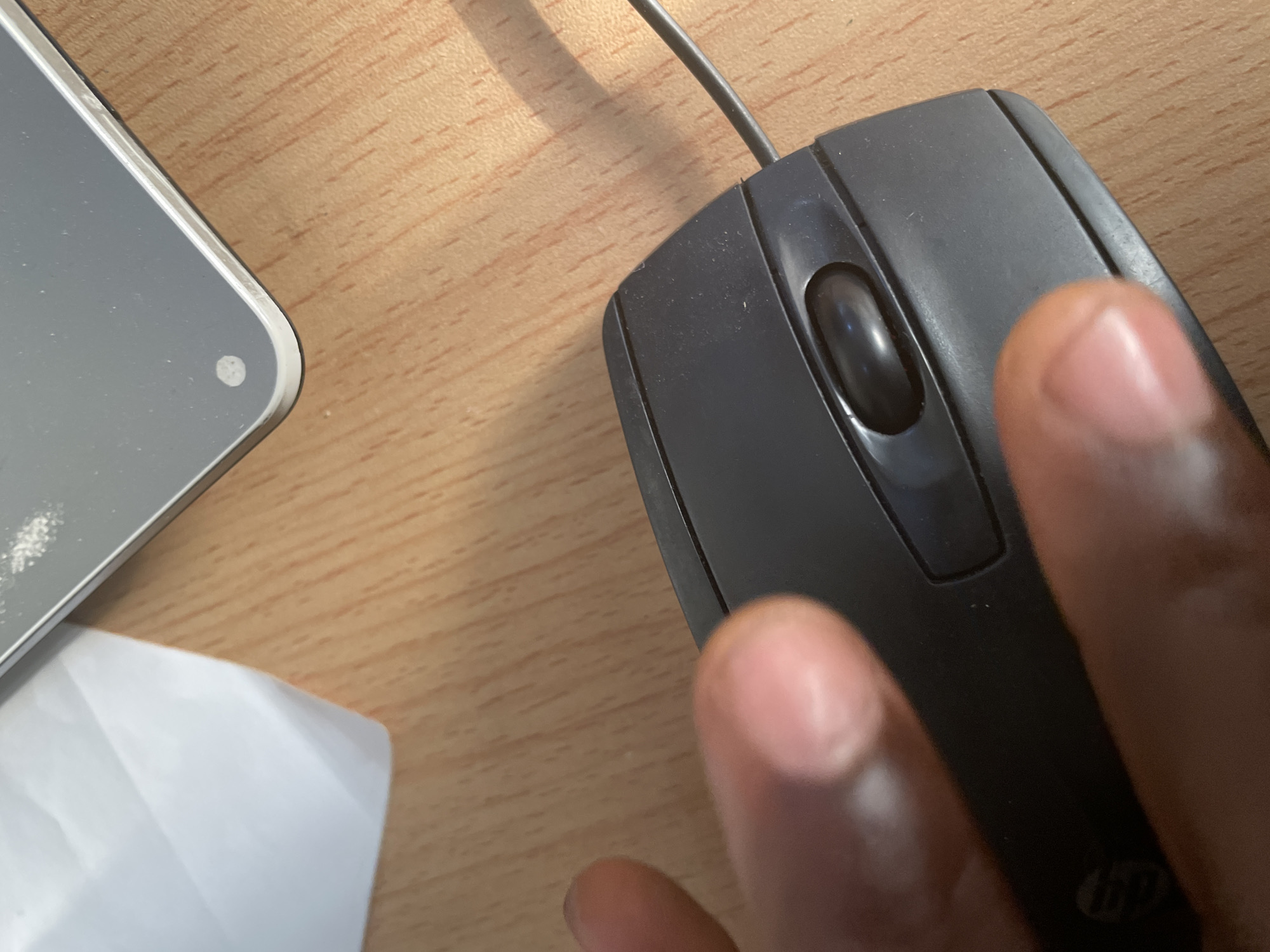 A close-up, overhead shot shows a dark gray computer mouse on a wooden desk. A person's hand, with dark skin, rests on the mouse, with fingers positioned over the buttons and scroll wheel. To the right of the mouse, a small corner of a silver electronic device is visible, with a smudge of white powder on its surface. A piece of white paper is also partially visible in the upper right quadrant of the image. The wooden desk surface exhibits a prominent wood grain pattern. The scene is static and lacks any discernible context of Jalingo, Nigeria.
