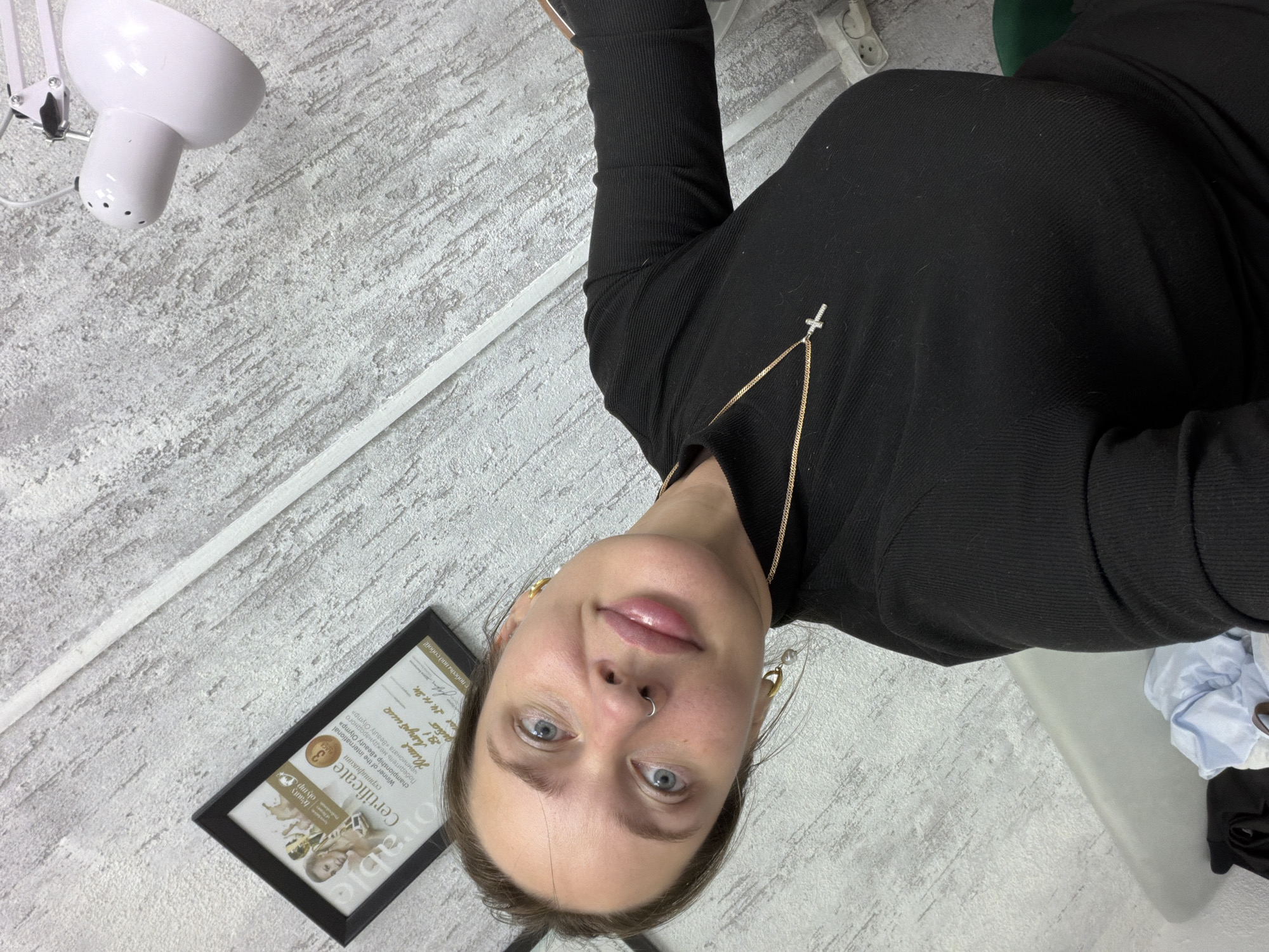 A young woman is depicted in a self-portrait, looking directly at the camera with a neutral to slightly smiling expression. She has blue eyes and a septum piercing, and wears a black, long-sleeved, ribbed top. Her attire is accessorized with a gold chain necklace featuring a small silver cross pendant, and gold earrings, one of which includes a pearl accent.
The scene appears to be an indoor professional or workspace, potentially a beauty salon or office located in Chaikovsky, Russia. The background features a light gray, textured wall or floor. In the upper left quadrant, a white, articulated desk lamp is visible. A prominent detail in the lower left is a framed certificate, clearly identifying the woman as "Юлия Ляпустина" (Yulia Lyapustina), the "Winner of the international championship «Beauty Olymp»," dated "24.11.24." The certificate also shows the word "LACE" and the phrase "победы над собой!" (victories over oneself!). Other elements in the background include a white power outlet, a white trim along the floor or wall, and some light blue fabric and dark objects suggesting a relaxed work setting. The lighting is artificial, characteristic of an indoor environment, making the time of day undeterminable. The woman's activity is posing for what appears to be a selfie.