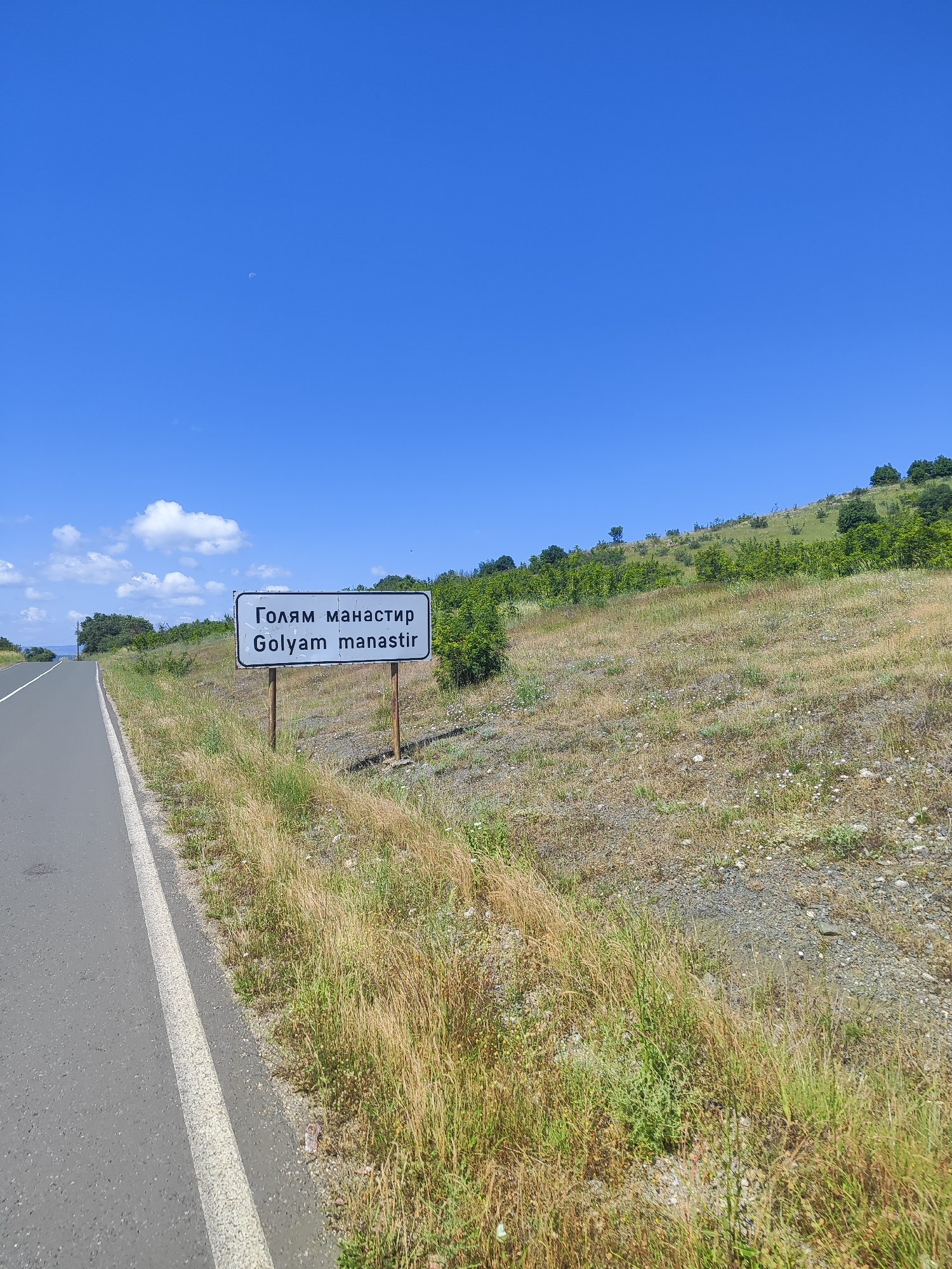 The image shows a sign with Bulgarian and Latin text indicating "Golyam Manastir". The sign is located next to a paved road on a dry, grassy hillside under a clear blue sky. The environment appears to be rural, with sparse vegetation and some trees on the distant hills. The sign is white with black text and is mounted on two wooden posts. The road has a white line marking the edge. The overall scene suggests a location in the countryside of Bulgaria, specifically the village of Golyam Manastir.