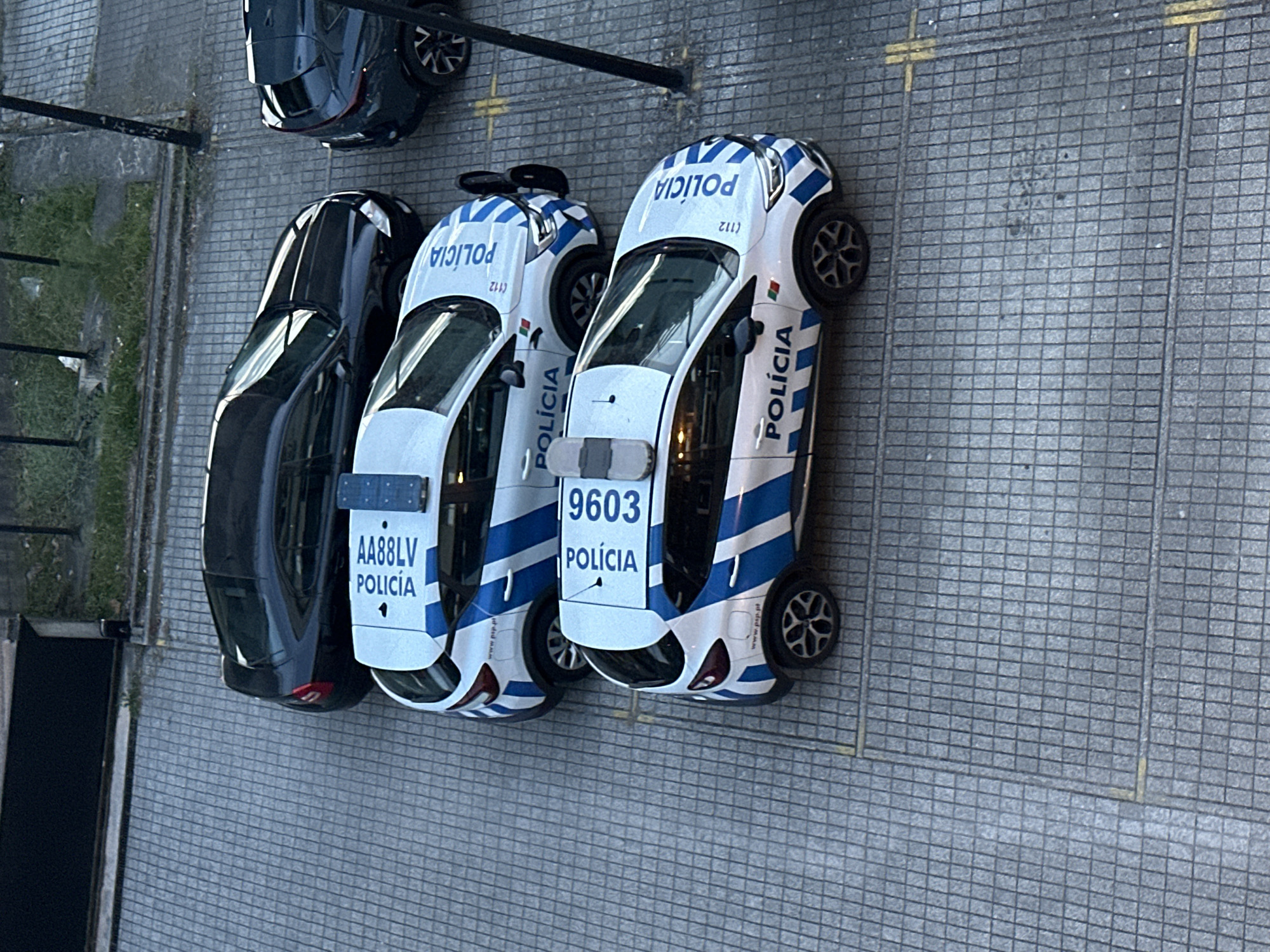 The image is a high-angle shot of three parked vehicles on a gray-tiled surface, with the top of the frame oriented towards the northeast. The vehicles are arranged in a line. On the left is a black passenger car. To the right are two white and blue police cars marked "POLÍCIA", which is the Portuguese word for police. The first police car has a license plate of "AA88LV". The second has the identification number "9603". A patch of grass is visible on the upper left of the frame and a black pole is visible just above the black car. The picture was taken in Vila Real, Portugal.