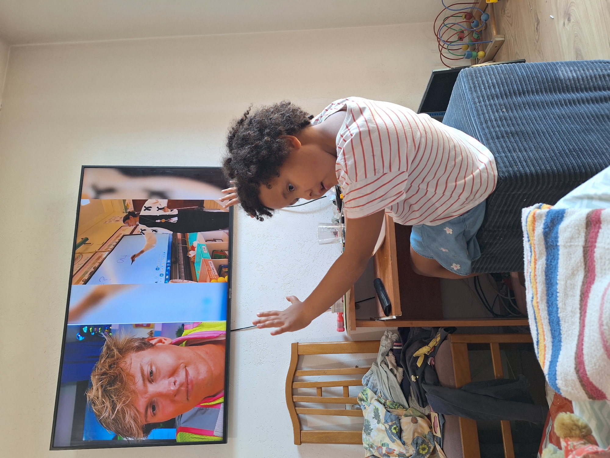 The image depicts an indoor scene, likely a living room or bedroom, with a young child positioned in the foreground. The child, with curly dark hair, is looking towards a large flat-screen television mounted on a white wall. The child is wearing a striped t-shirt and appears to be raising their right arm. The television displays a split screen. The left side shows a close-up of a young man with blonde, tousled hair, wearing a reflective vest. The right side shows a woman in front of a whiteboard, seemingly giving a lesson, with the number "94" written on the board.
In the background, a wooden chair is visible, partially covered with clothing. A small wooden desk or table is to the right of the chair, with a remote control and a glass on it. Further to the right, a colorful wooden bead maze toy is on the floor. The overall environment suggests a domestic setting. The presence of a television and a child interacting with it indicates an educational or entertainment context. There is no visible indication of the location being in Blagoevgrad, Bulgaria, or any other specific city.