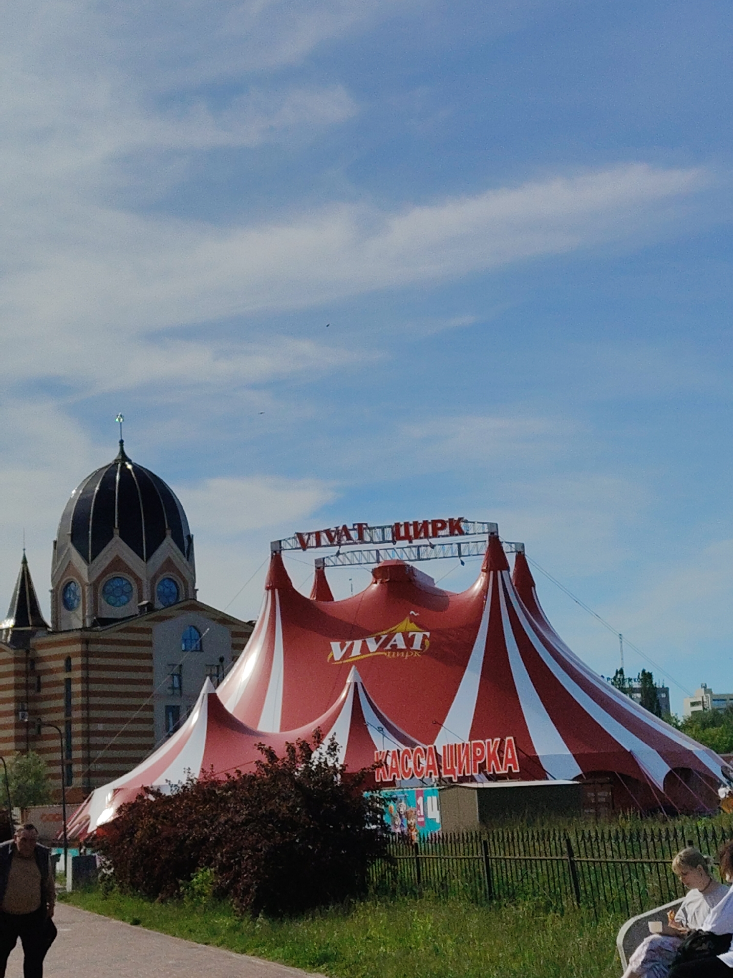 The image captures a red and white striped circus tent under a blue sky with scattered clouds. The tent is prominently branded with the word "VIVAT" and "ЦИРК" in large red letters on its peak and a smaller sign reading "КАССА ЦИРКА" (Circus Ticket Office) below. To the left of the tent stands a building with a distinctive dome and striped facade, resembling a synagogue. The building is made of tan brick with darker brown horizontal stripes and features several blue-domed windows. A smaller conical spire is visible on the left side of this building. In the foreground, a paved walkway is visible with a man walking away from the camera. To the right, a group of people are seated on benches in a grassy area, with one person eating. A dark green bush is situated in front of the circus tent. The overall scene suggests a public event or attraction in an urban setting. This circus tent is located at the Kaliningrad Circus.